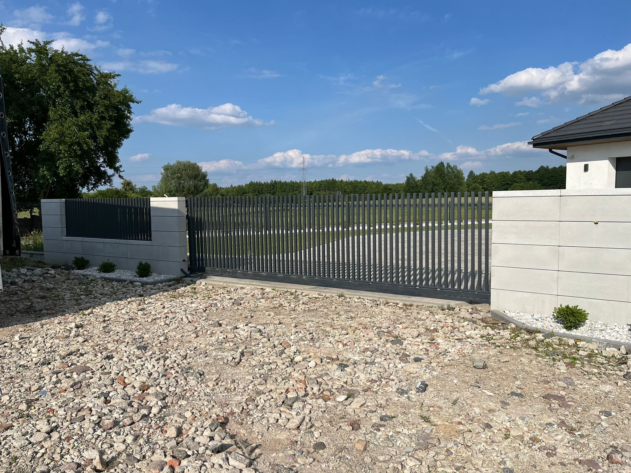 Nowoczesna, przesuwna brama z pionowych profili, osadzona między betonowymi słupkami wykończonymi drobnymi, białymi kamykami, na tle błękitnego nieba z lekkimi obłokami.