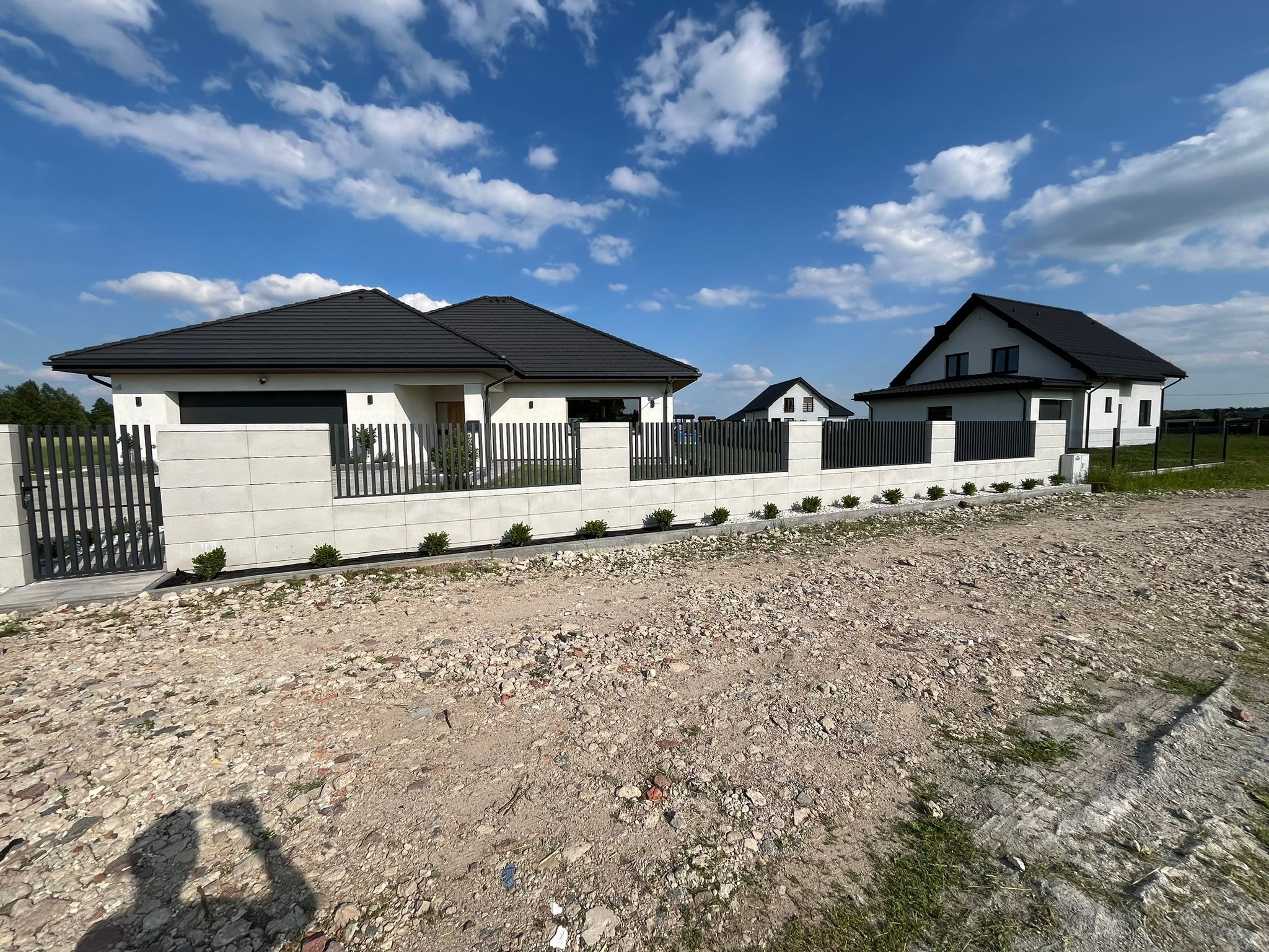 Nowoczesne ogrodzenie z metalowymi przęsłami i betonowymi słupkami, otaczające domy jednorodzinne w słoneczny dzień.