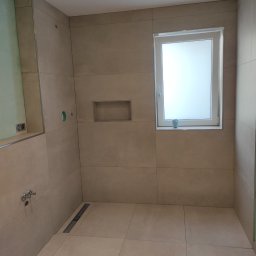 Usługi glazurnicze i remontowo budowlane JG - Beżowa łazienka z dużymi płytkami, odpływem liniowym, wnęką w ścianie i małym oknem z kubkiem na parapecie.