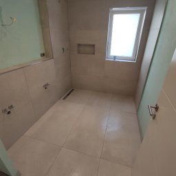 Usługi glazurnicze i remontowo budowlane JG - Wnętrze łazienki w trakcie wykończenia, z beżowymi płytkami, oknem dachowym i widocznymi instalacjami wodnymi przed montażem armatury.