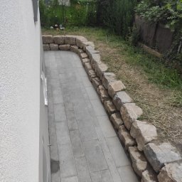 Usługi glazurnicze i remontowo budowlane JG - Ułożona szara kostka brukowa tworząca ścieżkę wzdłuż ściany budynku, ograniczona niskim murem z kamieni polnych, z trawnikiem w tle.
