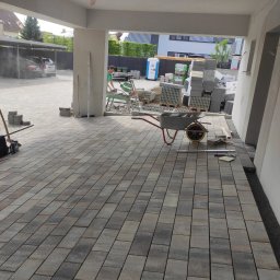 Usługi glazurnicze i remontowo budowlane JG - Nowo ułożona, prostokątna kostka brukowa w odcieniach szarości, z widocznym wózkiem budowlanym, paletami kostki i pracownikiem używającym piły do cięcia kostki w tle.