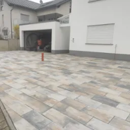 Ułożona kostka brukowa w odcieniach szarości, beżu i bieli na podjeździe przed nowoczesnym domem z garażem, widoczne narzędzia w garażu.