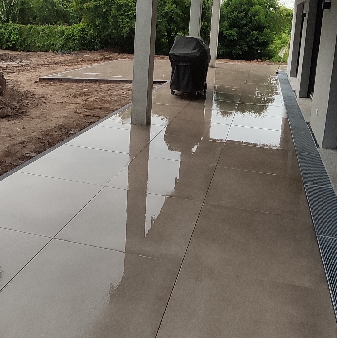 Wyłożony jasnoszarymi płytkami taras z widocznym grillem pod pokrowcem i fragmentem ogrodu z przygotowanym podłożem, po deszczu.