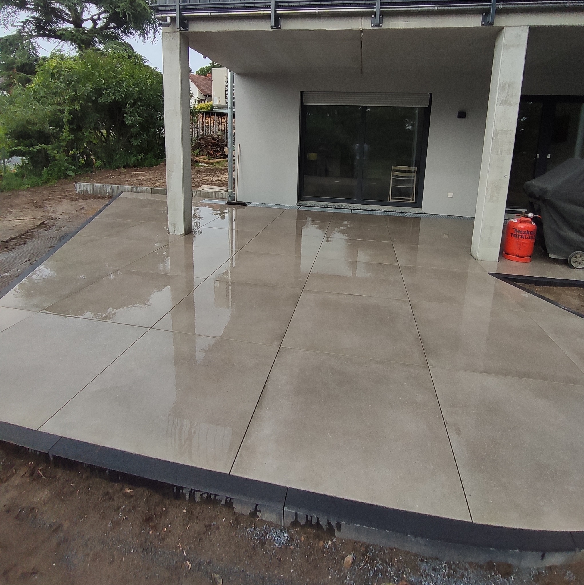 Świeżo ułożony, mokry taras z dużych, szarych płyt na zewnątrz budynku z widocznymi elementami ogrodu i butlą gazową w tle.