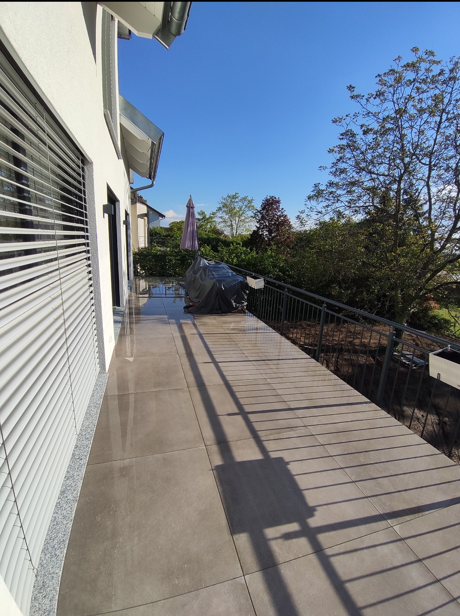 Balkon wyłożony szarymi płytkami ceramicznymi, z aluminiowymi żaluzjami na ścianie budynku i metalową barierką z doniczkami, widok na zieleń drzew w słoneczny dzień.