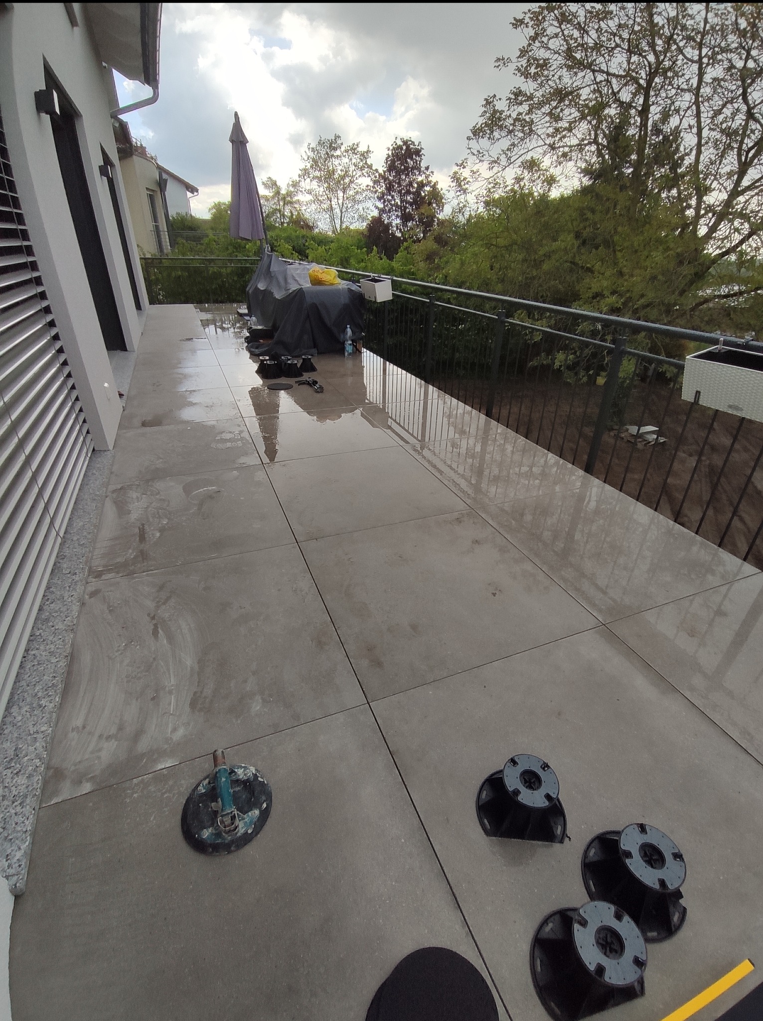 Montaż tarasu z płyt na regulowanych wspornikach; widoczne płytki, wsporniki, przyssawka do przenoszenia płytek i częściowo zadaszony balkon z metalową barierką w tle.