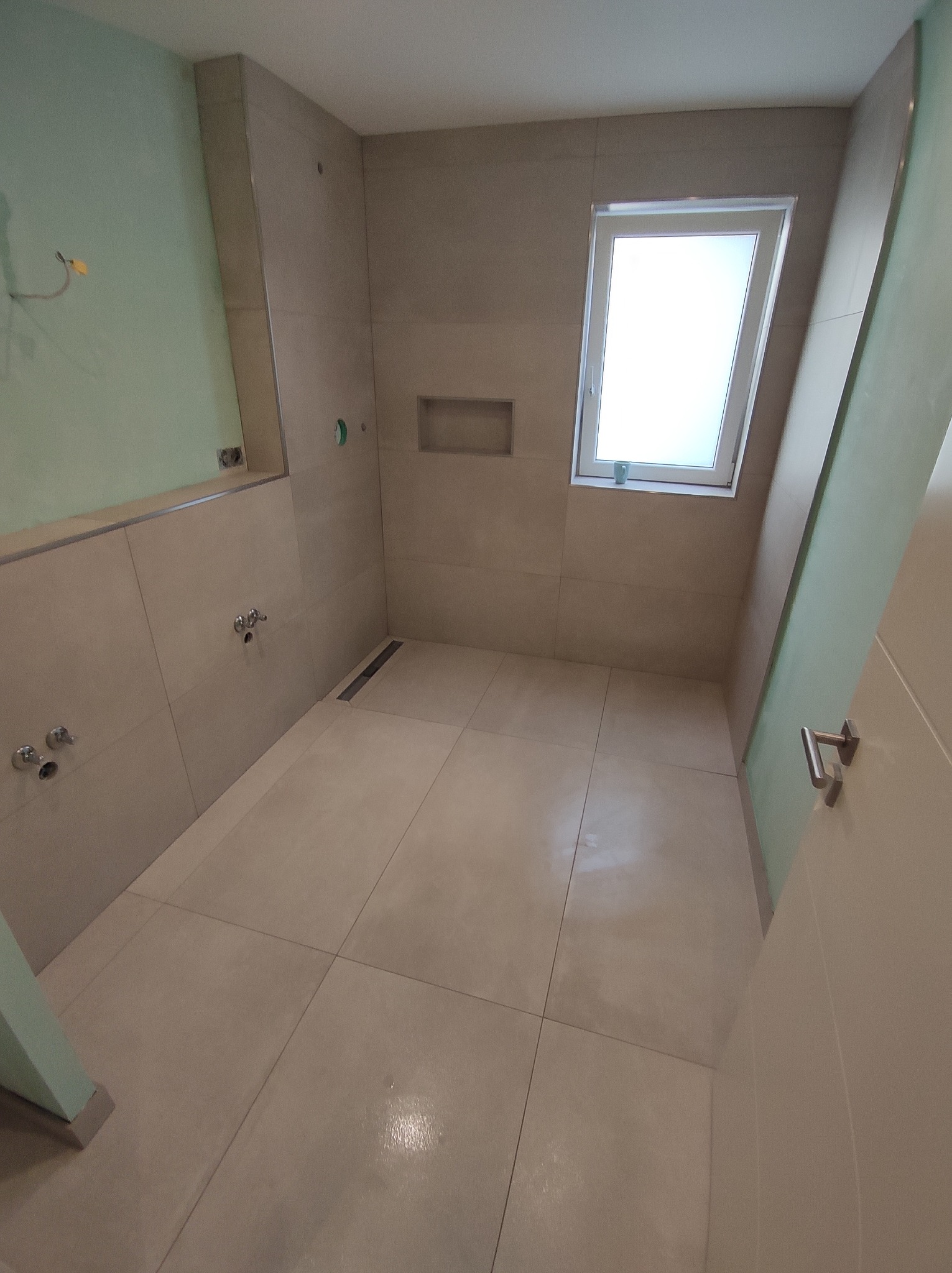 Wnętrze łazienki w trakcie wykończenia, z beżowymi płytkami, oknem dachowym i widocznymi instalacjami wodnymi przed montażem armatury.