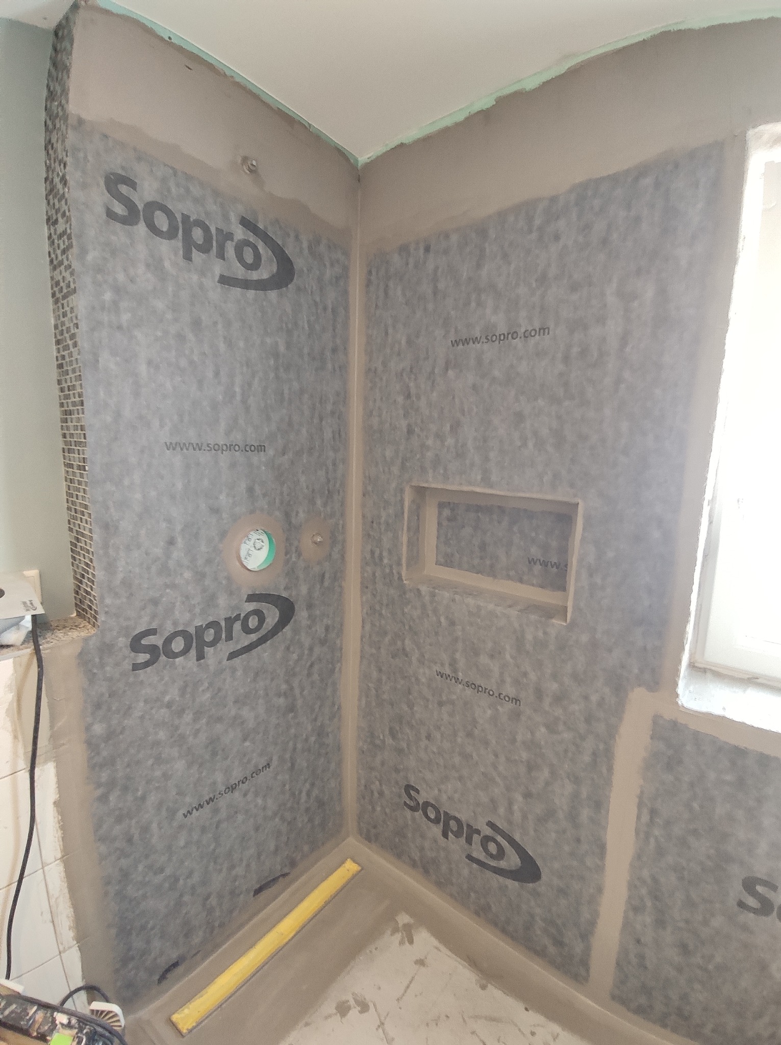 Przygotowanie ścian w kabinie prysznicowej do położenia płytek, widoczna warstwa hydroizolacji z logo Sopro, wnęka w ścianie, częściowo wykończona ściana mozaiką.