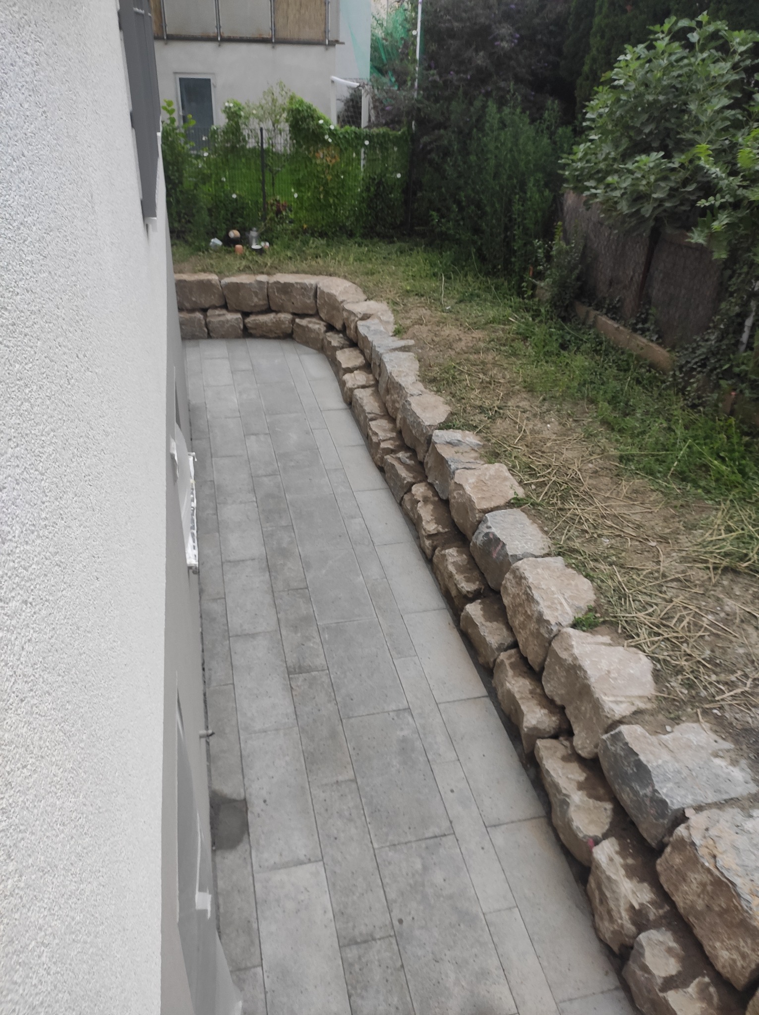 Ułożona szara kostka brukowa tworząca ścieżkę wzdłuż ściany budynku, ograniczona niskim murem z kamieni polnych, z trawnikiem w tle.