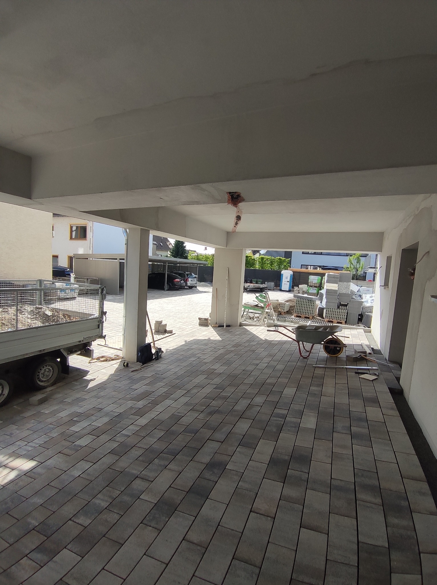 Ukończona nawierzchnia z szarej kostki brukowej pod zadaszeniem, widoczny wózek budowlany, przyczepa z materiałami i narzędzia, teren budowy.