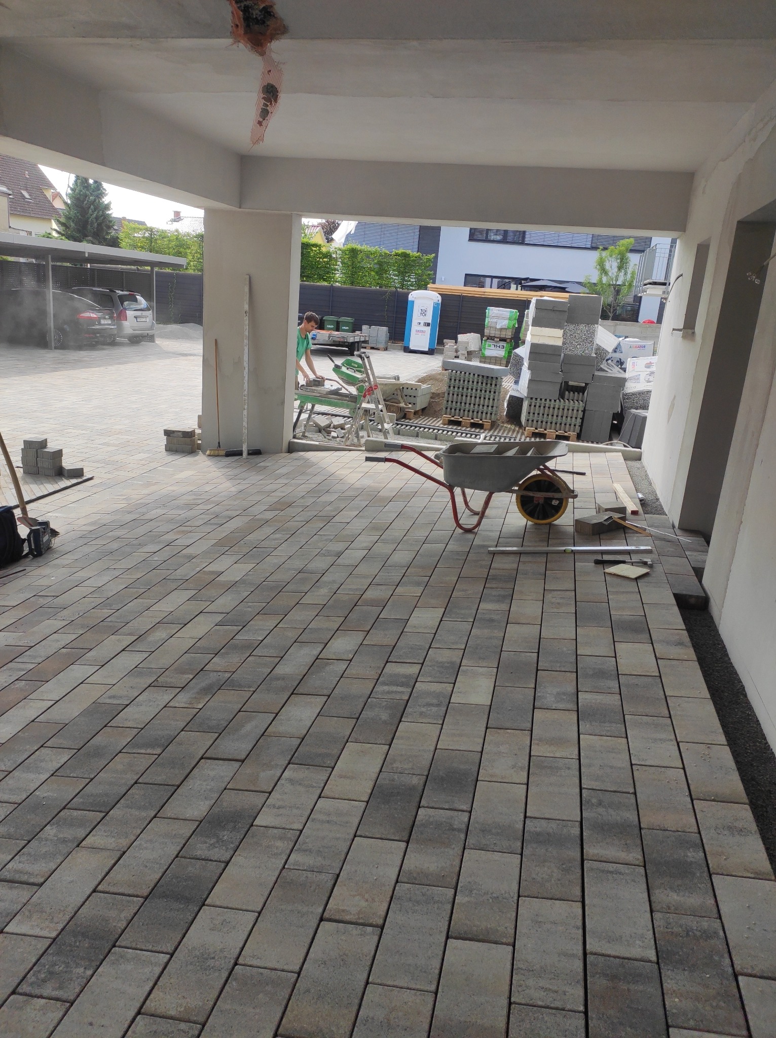 Nowo ułożona, prostokątna kostka brukowa w odcieniach szarości, z widocznym wózkiem budowlanym, paletami kostki i pracownikiem używającym piły do cięcia kostki w tle.