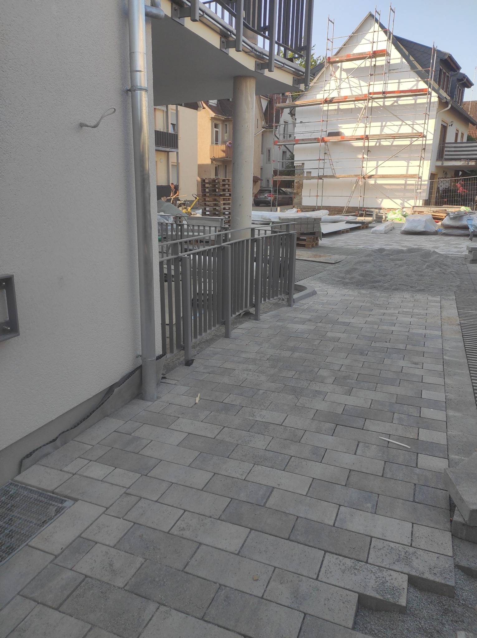 Ułożona szara kostka brukowa przed budynkiem z metalową balustradą, widoczny fragment elewacji budynku i materiały budowlane w tle.