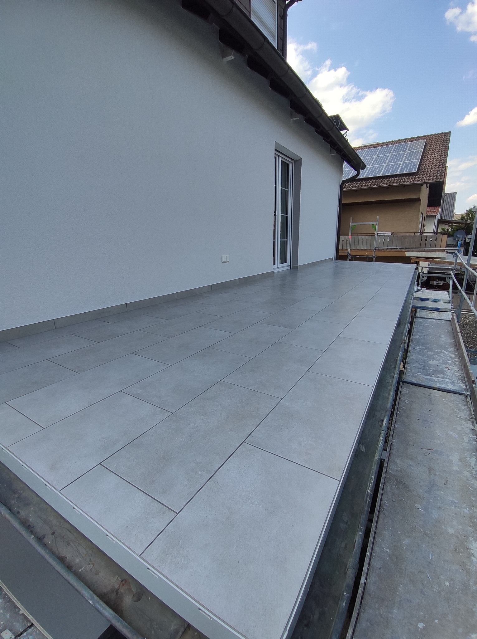 Wykończony taras z szarymi płytkami, widok z rusztowania, biała ściana budynku z drzwiami balkonowymi, w tle dom z panelami słonecznymi na dachu.