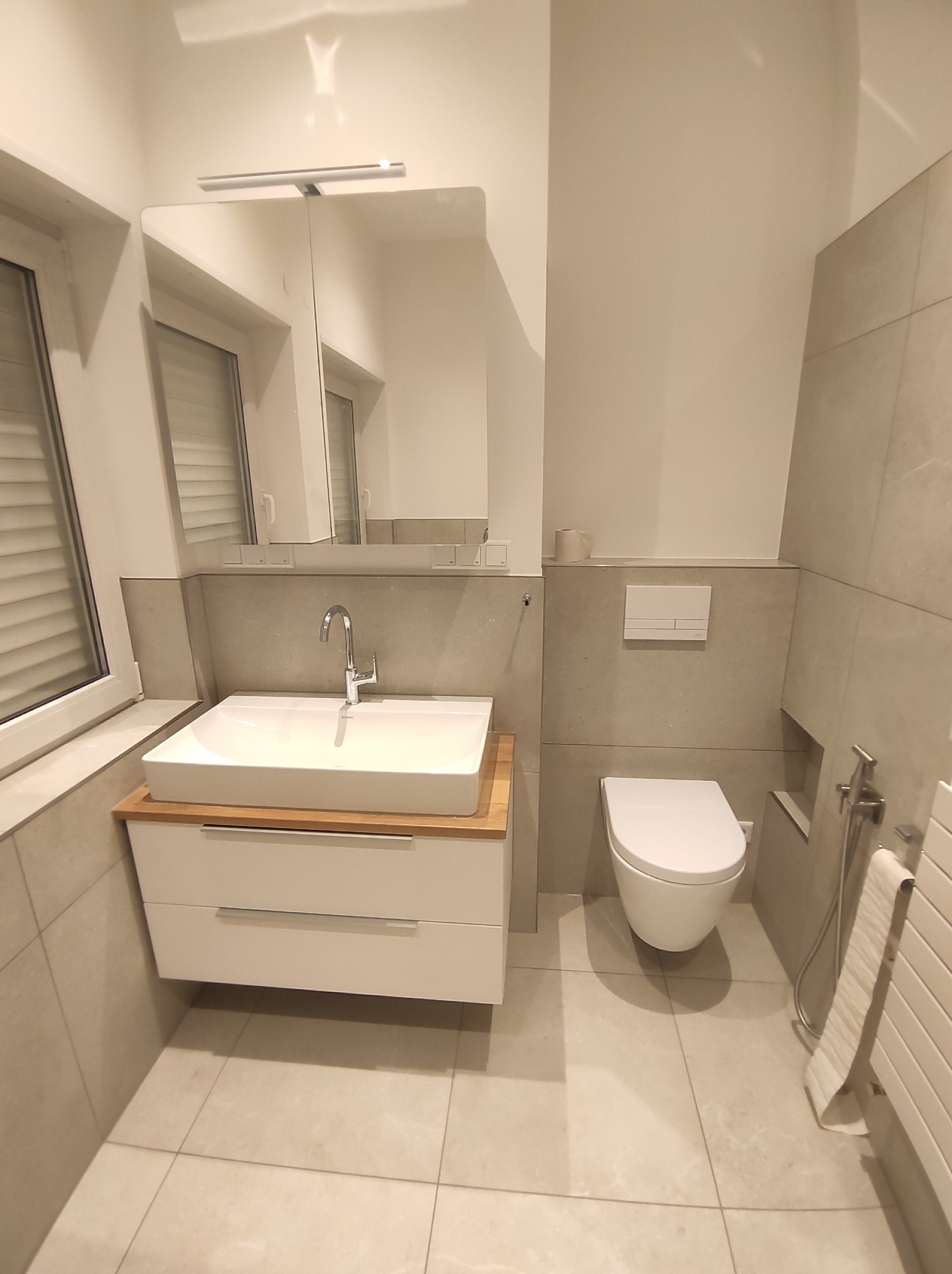 Nowoczesna łazienka w odcieniach szarości i bieli, z minimalistyczną umywalką nablatową na drewnianym blacie, podświetlanym lustrem i wiszącą miską WC.