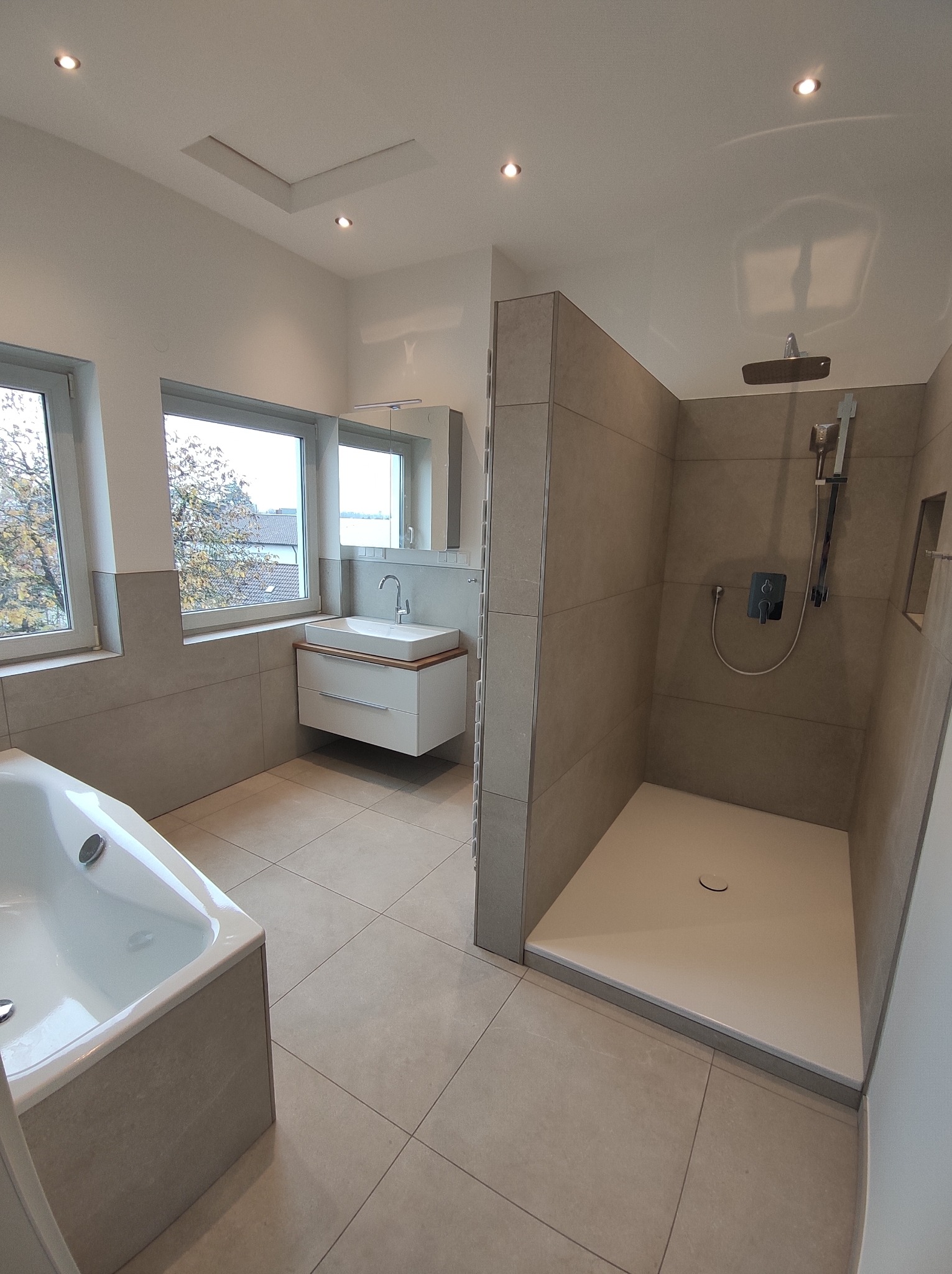 Nowoczesna łazienka z wanną, umywalką z szafką i kabiną prysznicową typu walk-in, wykończona płytkami w odcieniach szarości, z oknem i oświetleniem punktowym.