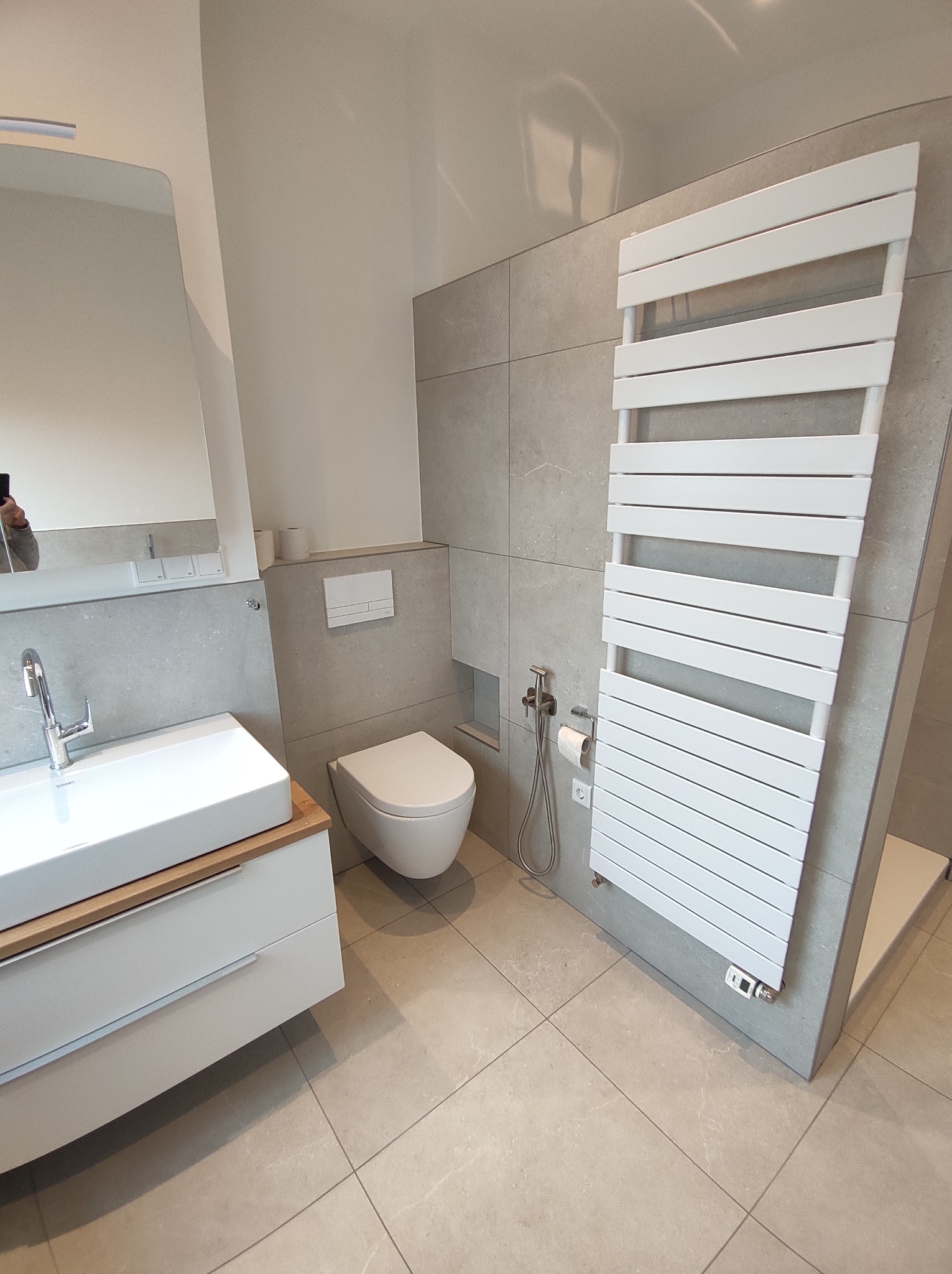 Nowoczesna łazienka w odcieniach szarości z białymi elementami: umywalka nablatowa z drewnianym blatem, podwieszana toaleta, biały grzejnik drabinkowy i bidetka.