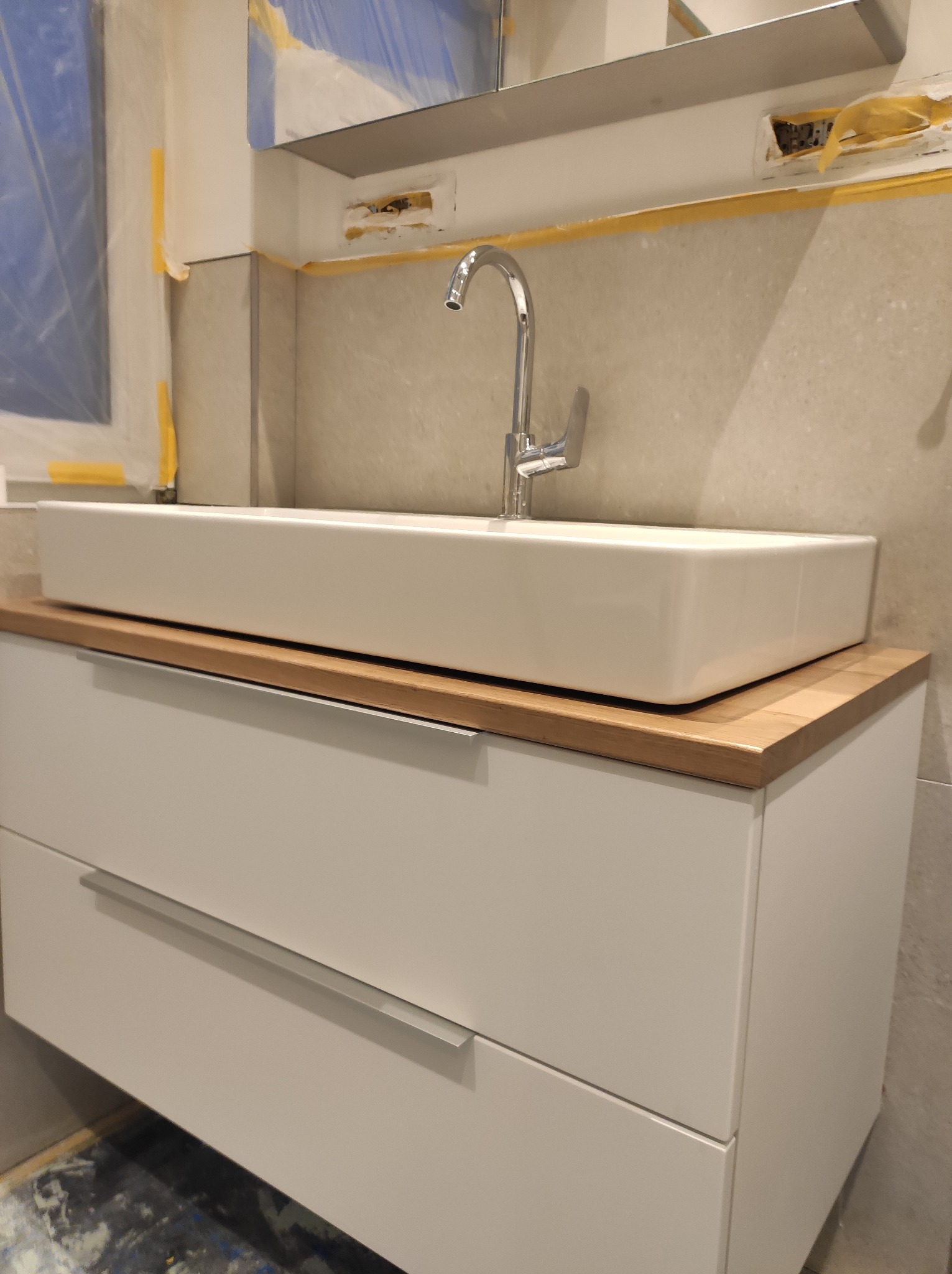 Nowoczesna, prostokątna umywalka nablatowa z chromowaną baterią, osadzona na szafce z dwiema szufladami z drewnianym blatem, widoczne zabezpieczenia folią malarską na ścianie i niedokończone prace...