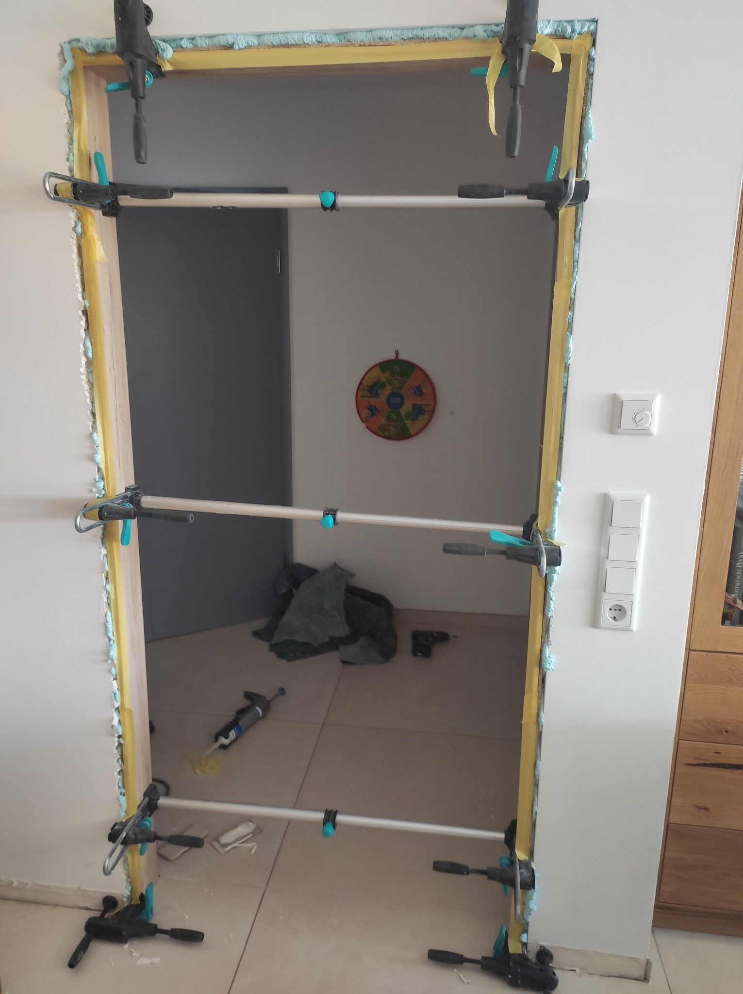 Montaż ościeżnicy drzwiowej z użyciem rozpórek teleskopowych, widoczna piana montażowa i taśma malarska zabezpieczająca ścianę, w tle widoczny cel do gry w rzutki.