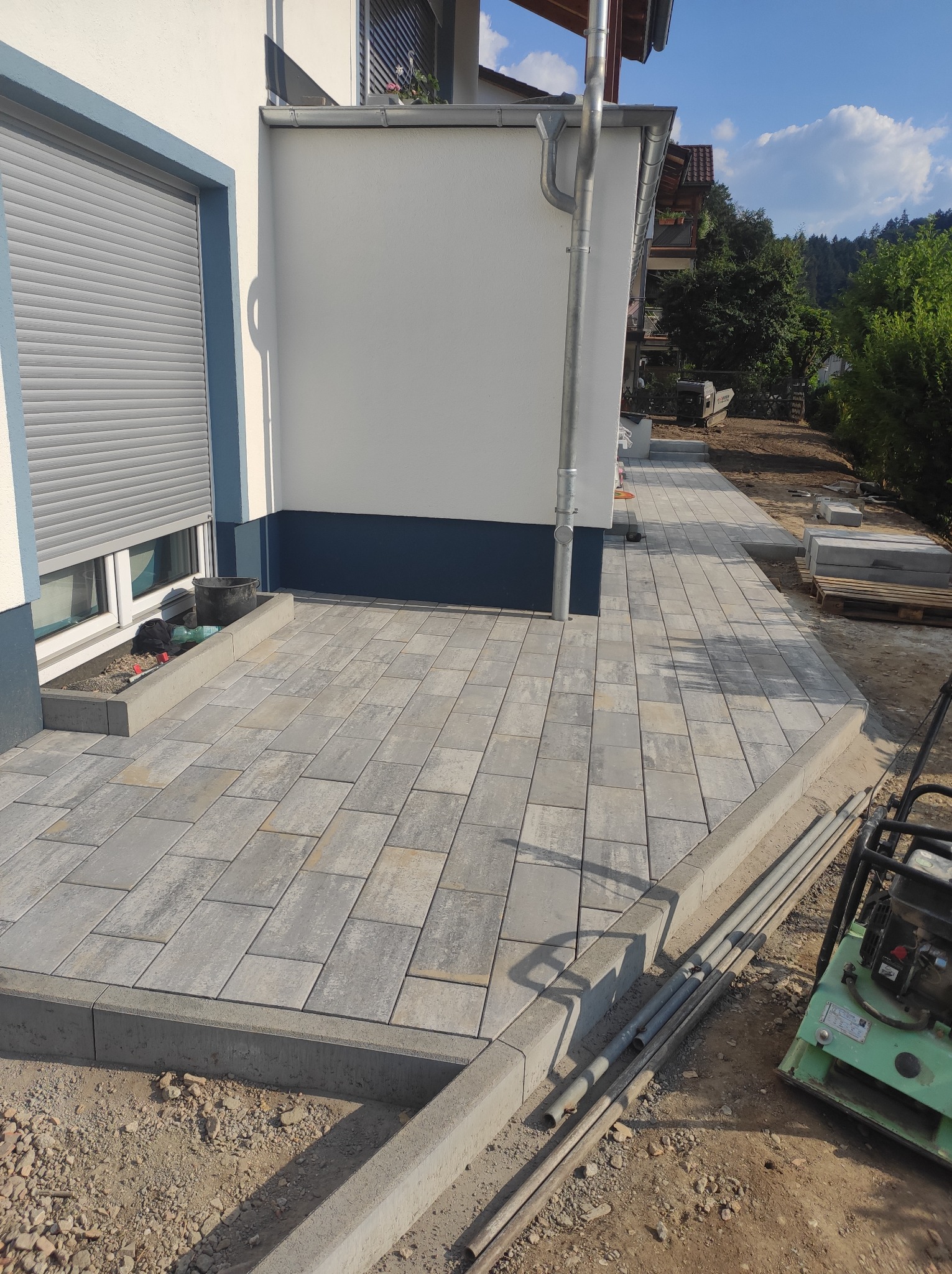 Świeżo ułożona, szara kostka brukowa na podwórku domu jednorodzinnego, z widoczną ubijarką wibracyjną i rurami na ziemi, w tle budynek z aluminiową żaluzją.