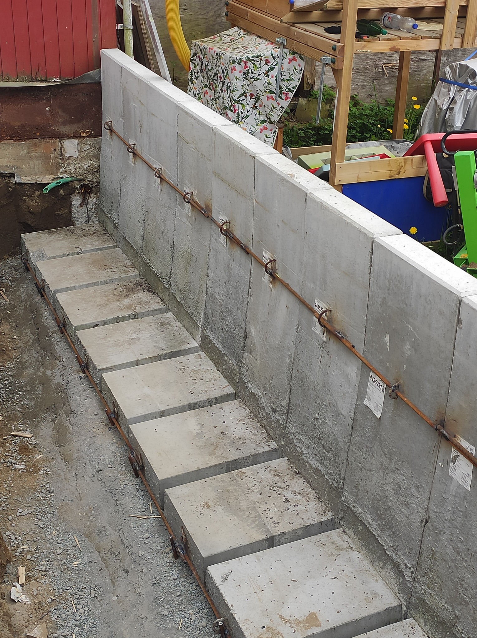 Wznoszenie betonowego muru oporowego z widocznymi elementami zbrojenia i etykietami na bloczkach, w tle elementy zagospodarowania terenu.