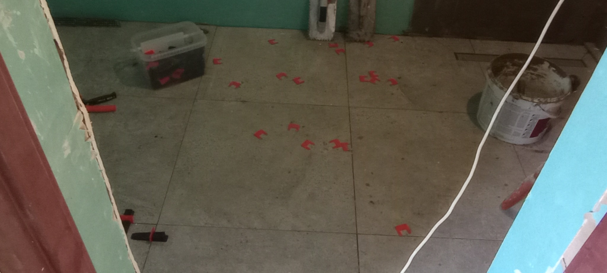 Fragment podłogi z szarych płytek, w trakcie układania, z widocznymi czerwonymi klipsami poziomującymi, pojemnikiem z klipsami, kielnią i brudnym wiadrem po kleju.