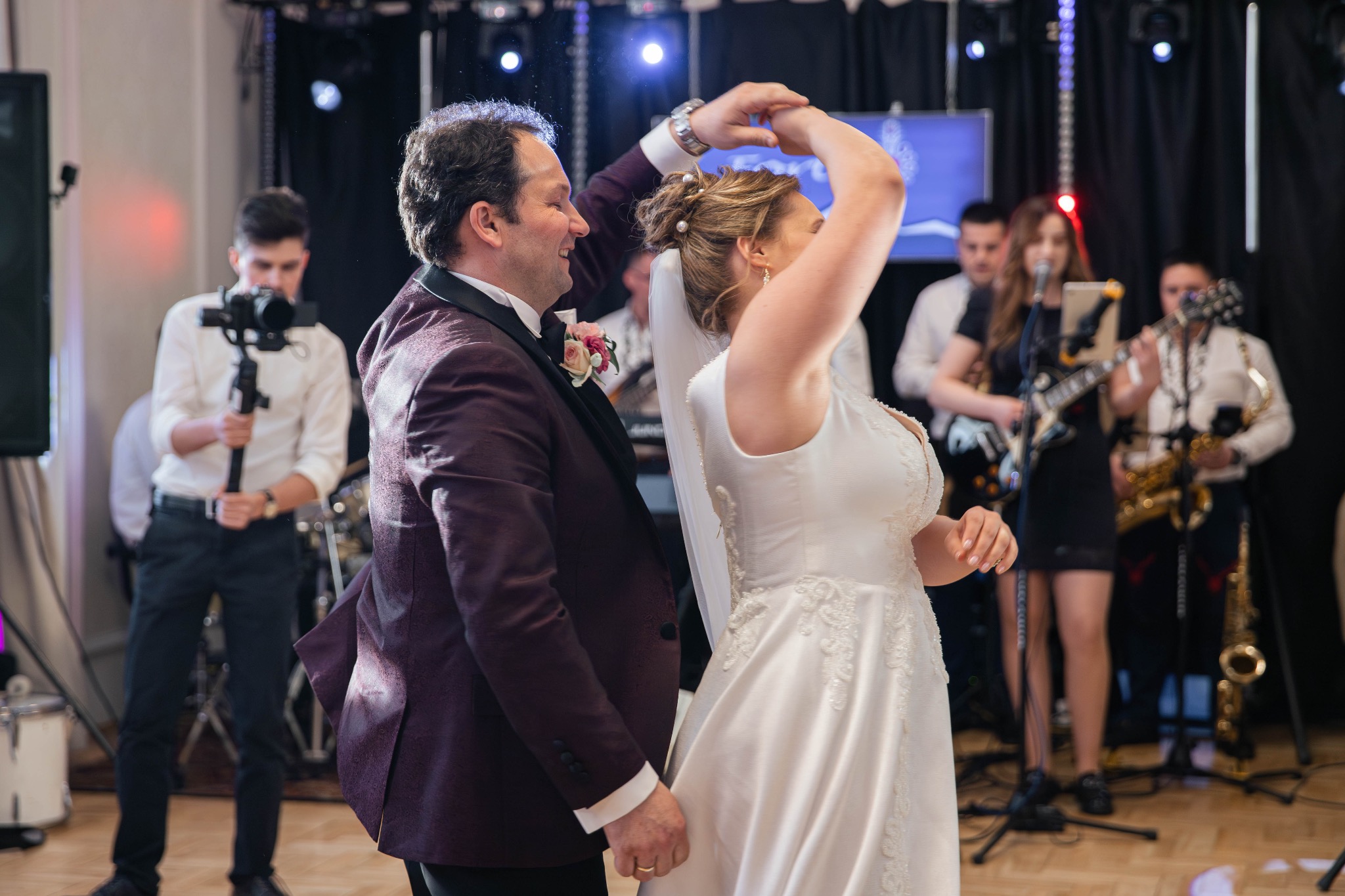 Para młoda w tańcu, on w bordowym smokingu, ona w białej sukni ślubnej, w tle zespół muzyczny i operator kamery.