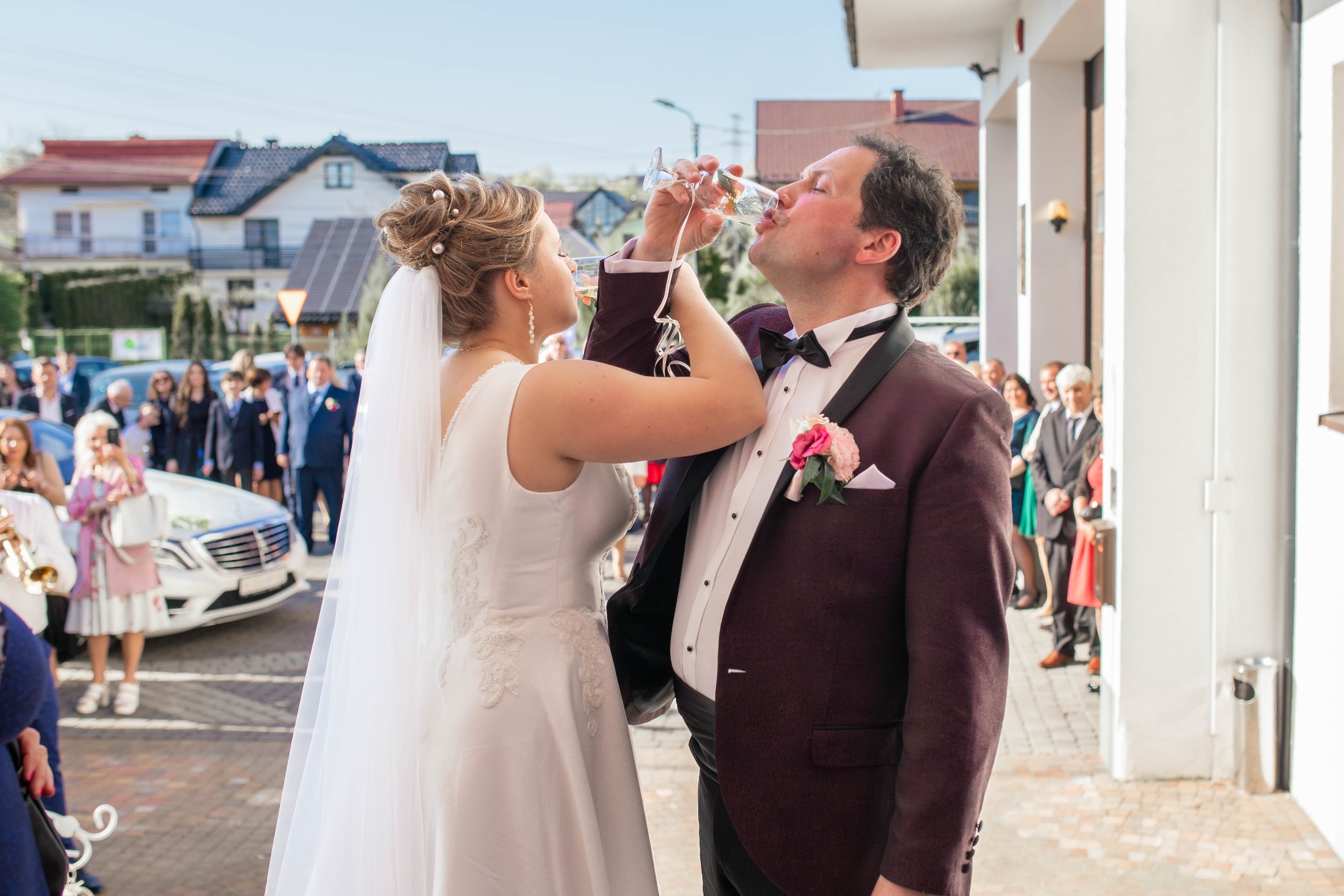 Para młoda w strojach ślubnych pije szampana z kieliszków na tle gości weselnych i białej limuzyny.