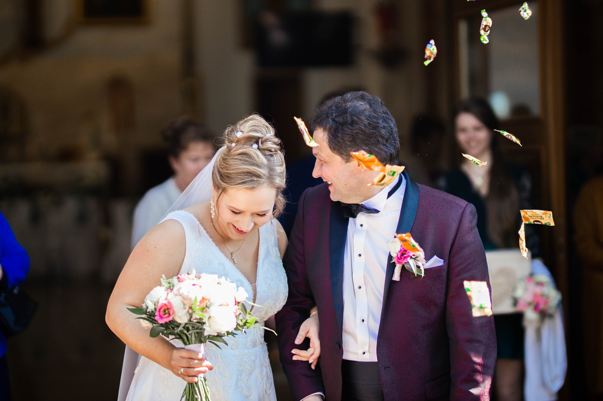 Uśmiechnięta para młoda wychodzi z budynku, obsypywana słodyczami w papierkach. Panna młoda w białej sukni trzyma bukiet, pan młody w bordowym garniturze z czarnymi klapami.