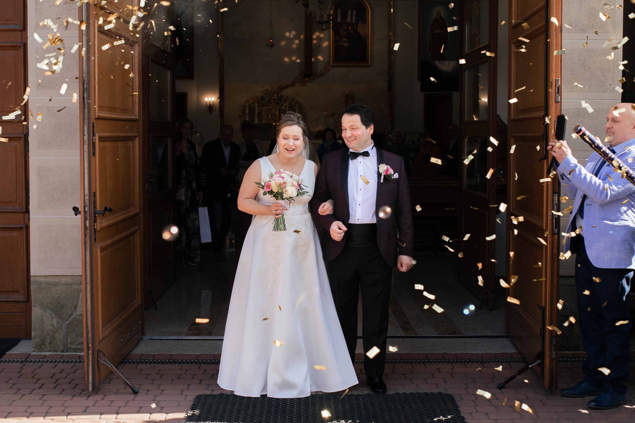 Para młoda, wychodząca z kościoła w Rudzie Śląskiej, obsypywana konfetti, z uśmiechami na twarzach i bukietem w ręku panny młodej, w tle widoczni goście.