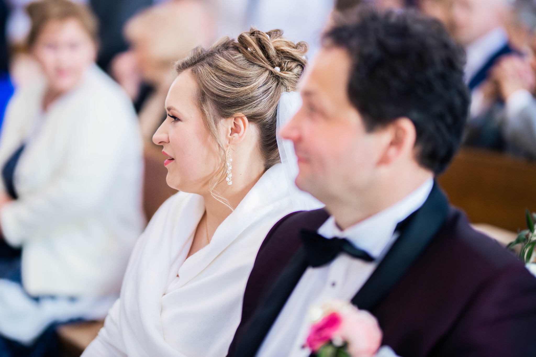 Para młoda siedząca obok siebie podczas uroczystości. Panna młoda ma upięte włosy z perłami i długie kolczyki, ubrana jest w białą narzutkę. Pan młody ma muchę i ciemny garnitur z butonierką.
