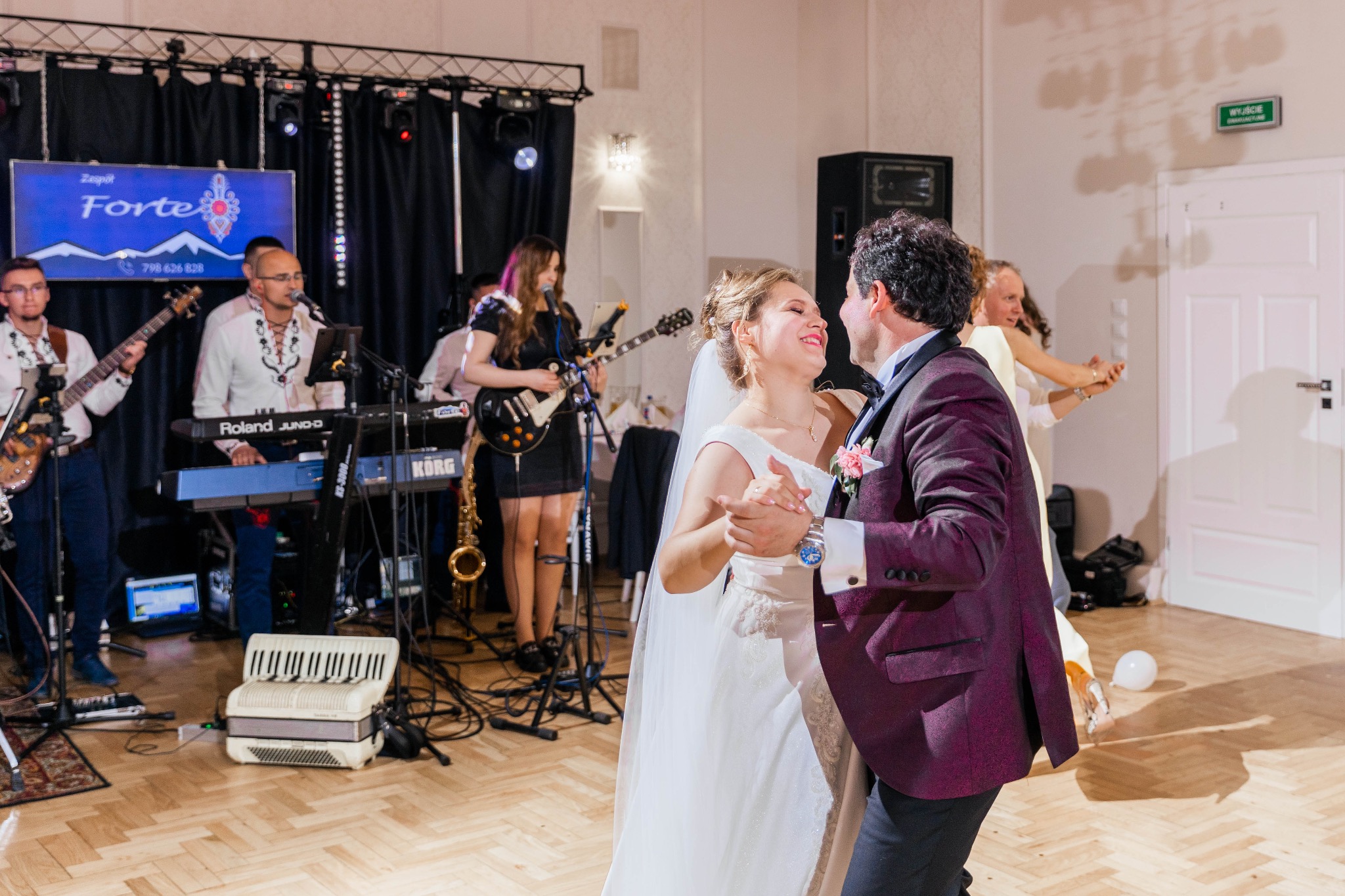 Para młoda w tańcu podczas wesela, w tle zespół muzyczny z akordeonem, gitarami i saksofonem, oświetlenie sceniczne.