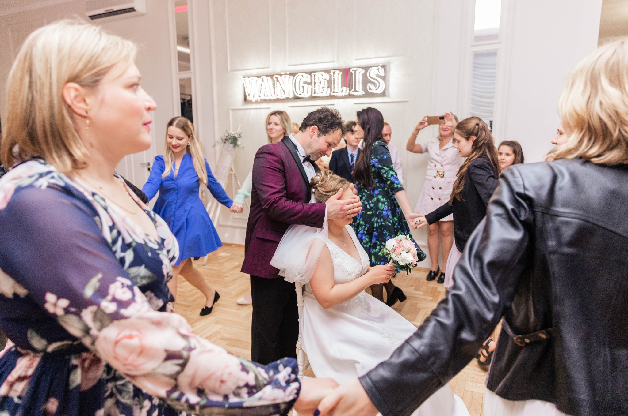 Para młoda podczas oczepin, otoczona gośćmi trzymającymi się za ręce, w tle neon z napisem 'VANGELIS'.