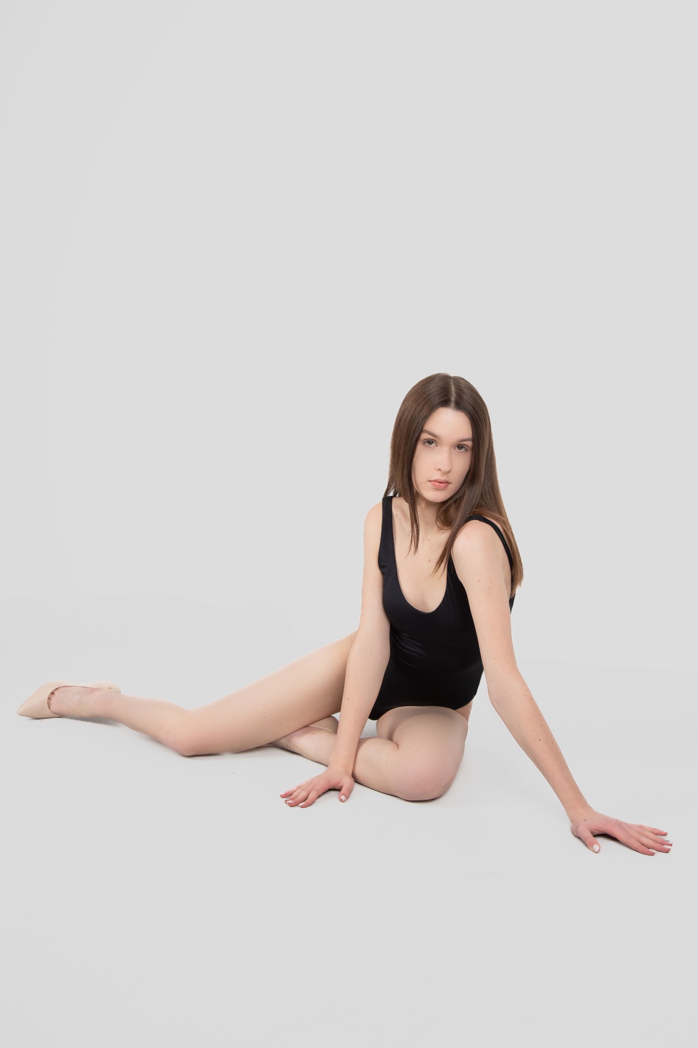 Kobieta w czarnym stroju kąpielowym i beżowych baletkach pozuje na jasnoszarym tle, siedząc na podłodze z wyciągniętą ręką.