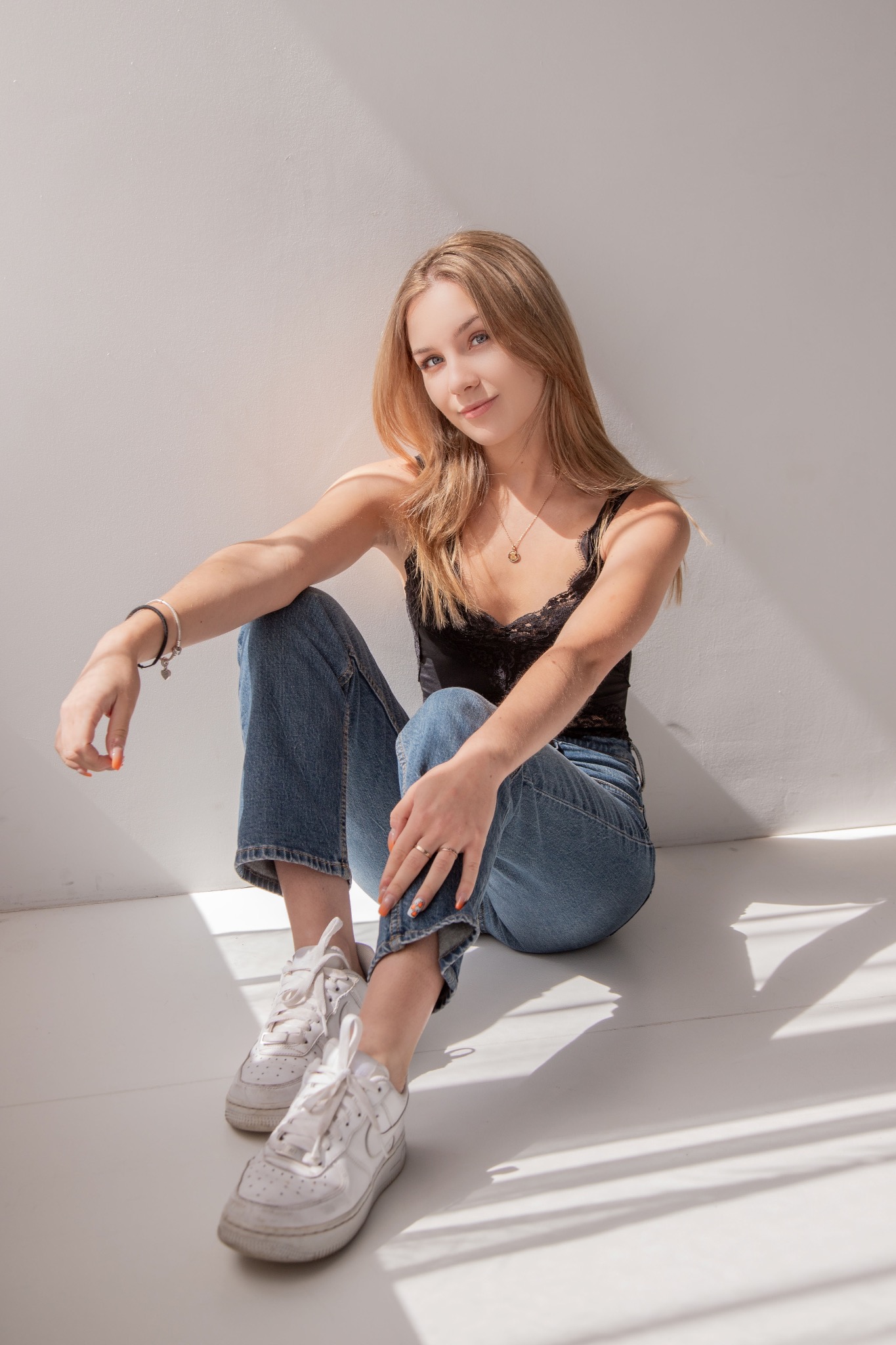 Portret młodej kobiety w jeansach i topie, siedzącej na podłodze w jasnym świetle, z delikatnym uśmiechem i subtelną biżuterią. Minimalistyczne tło.