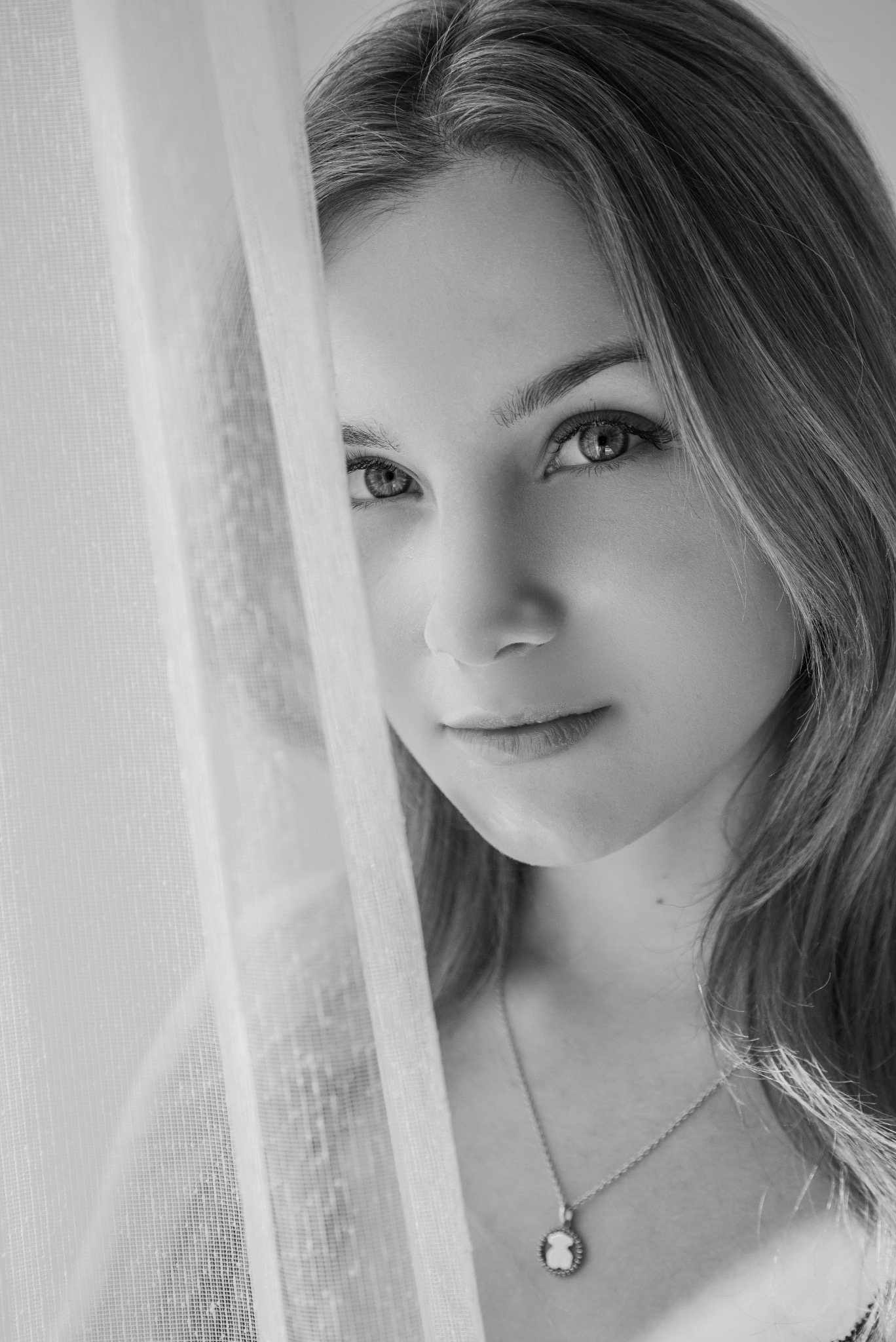 Portret młodej kobiety w czerni i bieli, z delikatną siatką firanki w tle, podkreślający jej spojrzenie i subtelny naszyjnik z misiem.