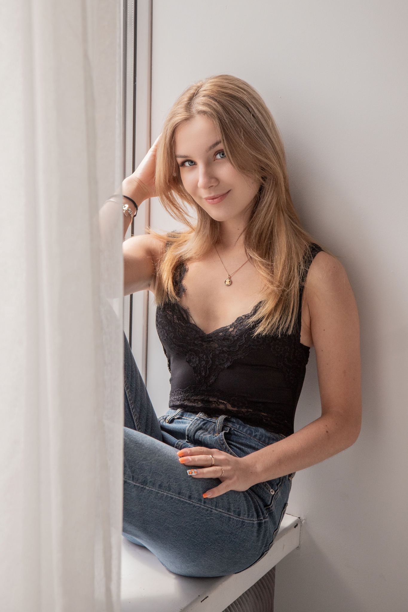 Portret uśmiechniętej blondynki w czarnym topie z koronką i jeansach, siedzącej przy oknie z ręką na włosach, patrzącej w obiektyw.