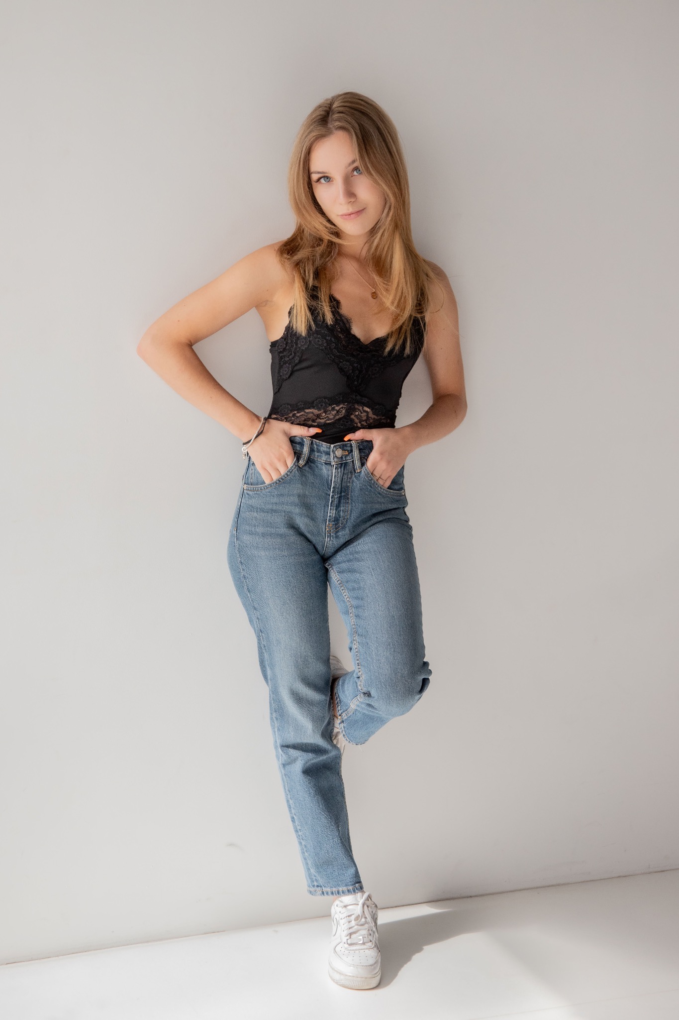Portret kobiety w dżinsach i czarnym topie z koronką, oparta o białą ścianę, z rękami w kieszeniach, w naturalnym świetle dziennym. Minimalistyczny styl.