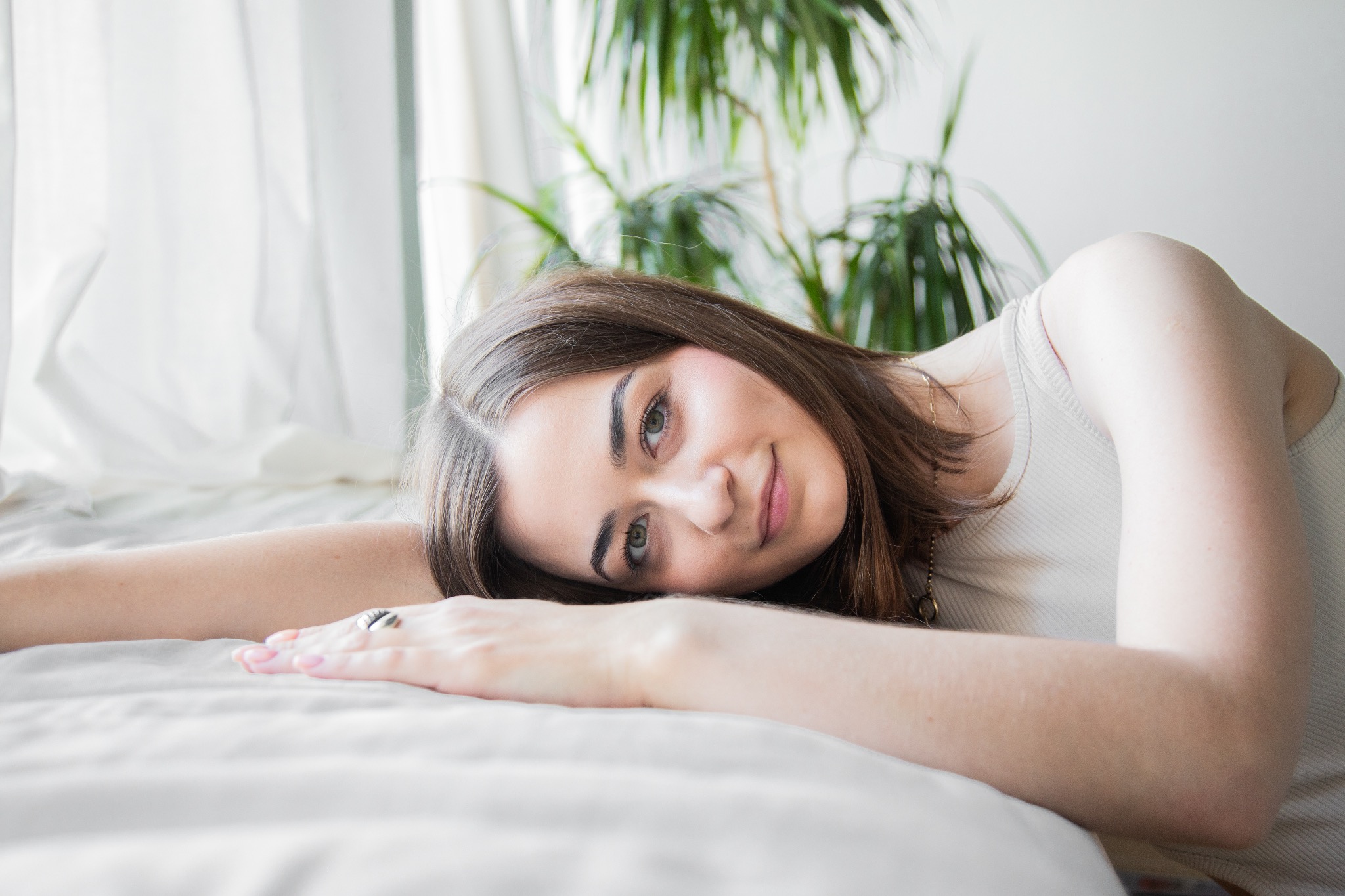 Portret uśmiechniętej brunetki leżącej na białej pościeli, z widocznym pierścionkiem na palcu, w tle okno z białymi zasłonami i zielona roślina doniczkowa.
