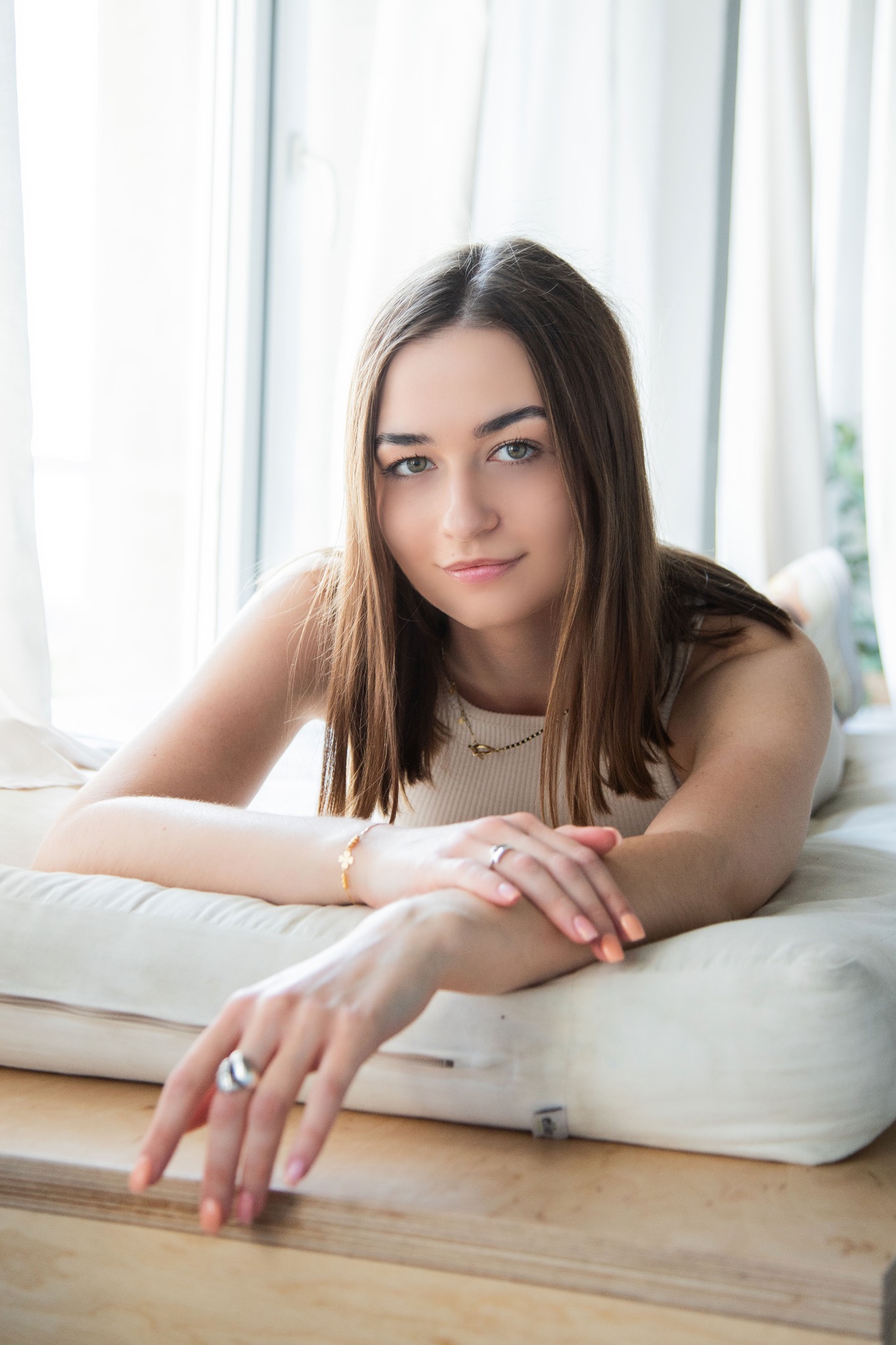 Portret młodej kobiety leżącej na jasnej poduszce przy oknie z białymi zasłonami, z delikatnym makijażem i biżuterią, w naturalnym świetle.