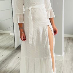 Anna's Fashion - Kobieta prezentuje długą, białą sukienkę z rozcięciem na nodze, wykonaną z delikatnego, prześwitującego materiału w drobne kropki, z rękawami 3/4 zakończonymi falbanami i wiązaniem w pasie.