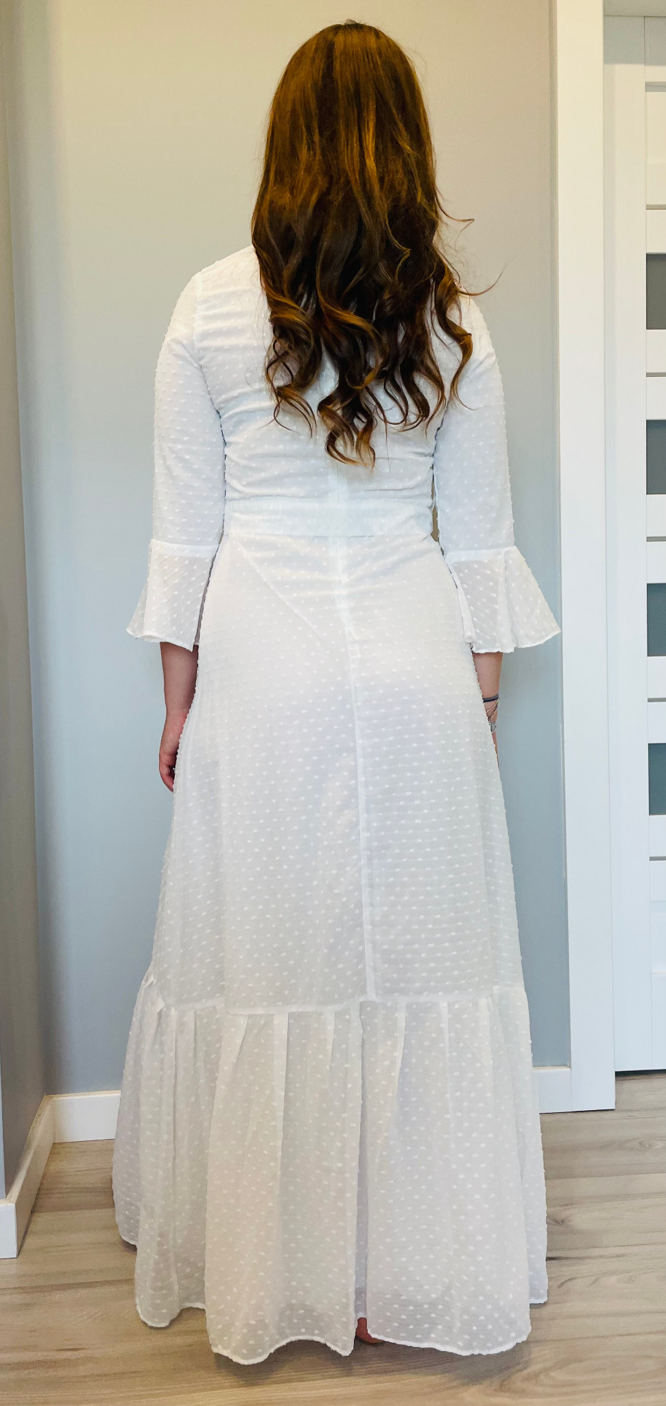 Kobieta z tyłu w długiej, białej, zwiewnej sukience z rękawami 3/4 wykończonymi falbanami, materiał w drobne kropki, brązowe kręcone włosy.