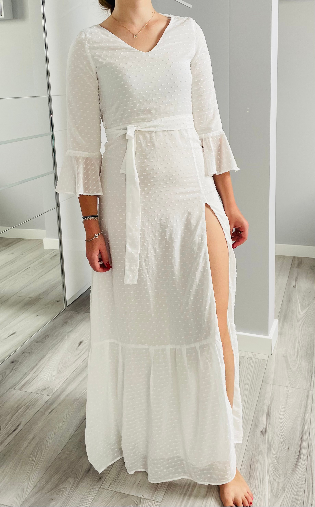 Kobieta prezentuje długą, białą sukienkę z rozcięciem na nodze, wykonaną z delikatnego, prześwitującego materiału w drobne kropki, z rękawami 3/4 zakończonymi falbanami i wiązaniem w pasie.