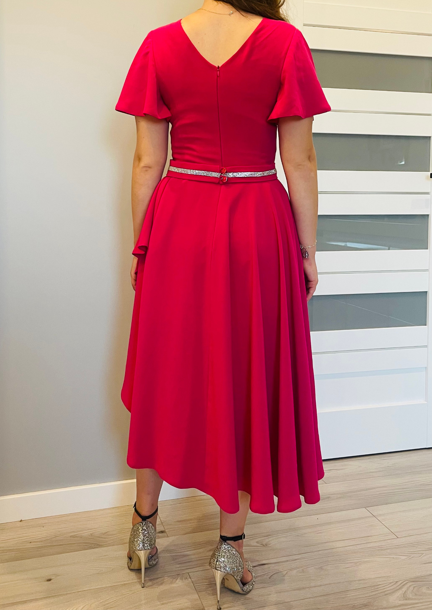 Kobieta w fuksjowej sukience z asymetrycznym dołem, ozdobnym paskiem i brokatowych szpilkach, widziana od tyłu na tle białych drzwi.