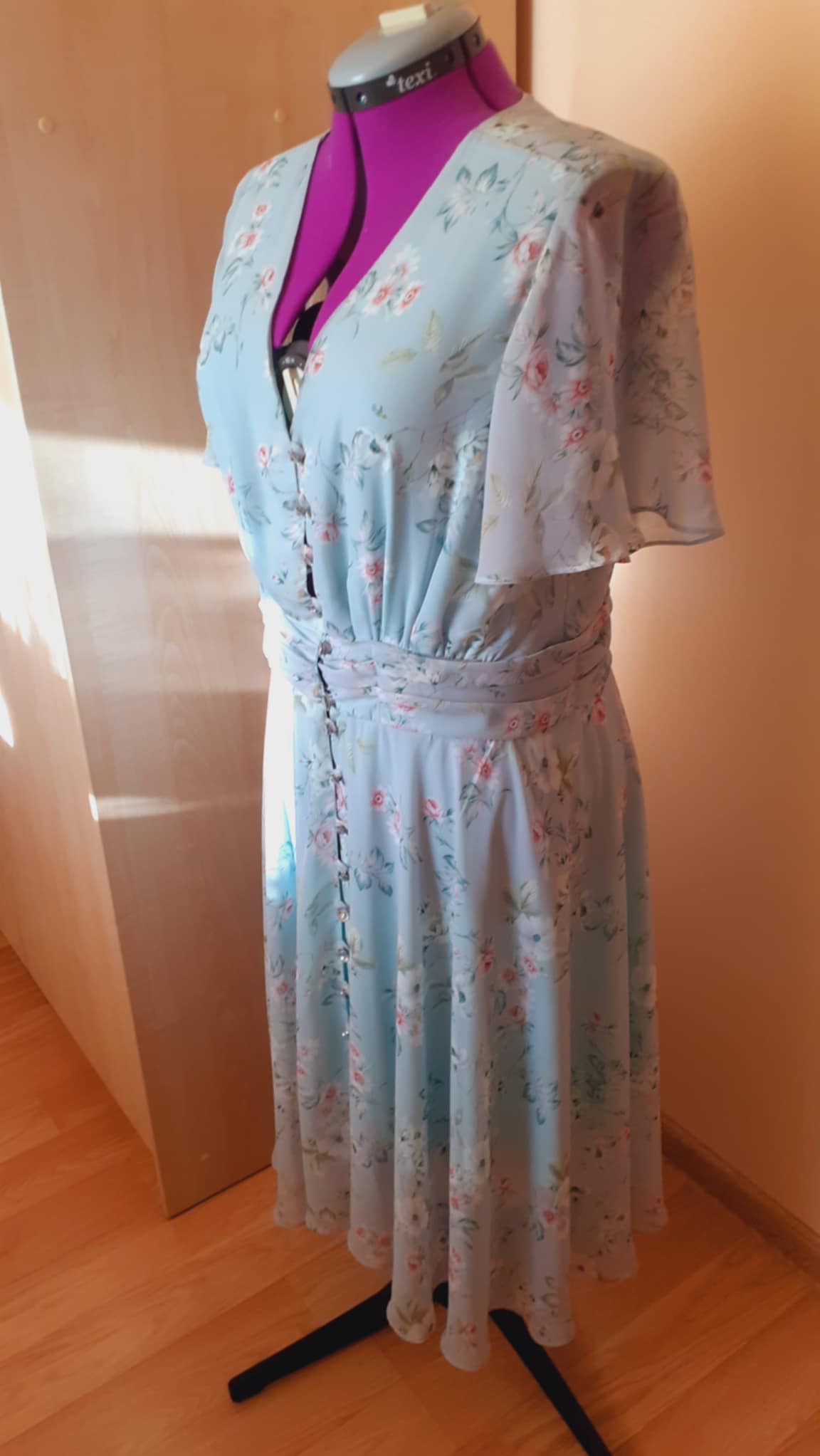 Jasnoniebieska, zwiewna sukienka w kwiaty na różowym manekinie krawieckim, widoczne guziki i detale wykończenia.