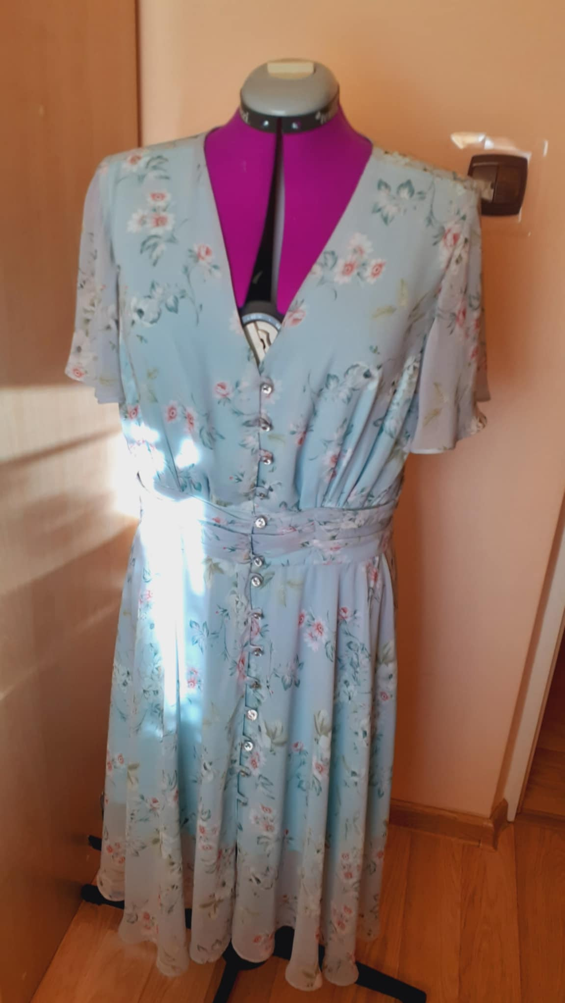 Jasnoniebieska, zwiewna sukienka w kwiaty, zapinana na guziki, prezentowana na różowym manekinie krawieckim.