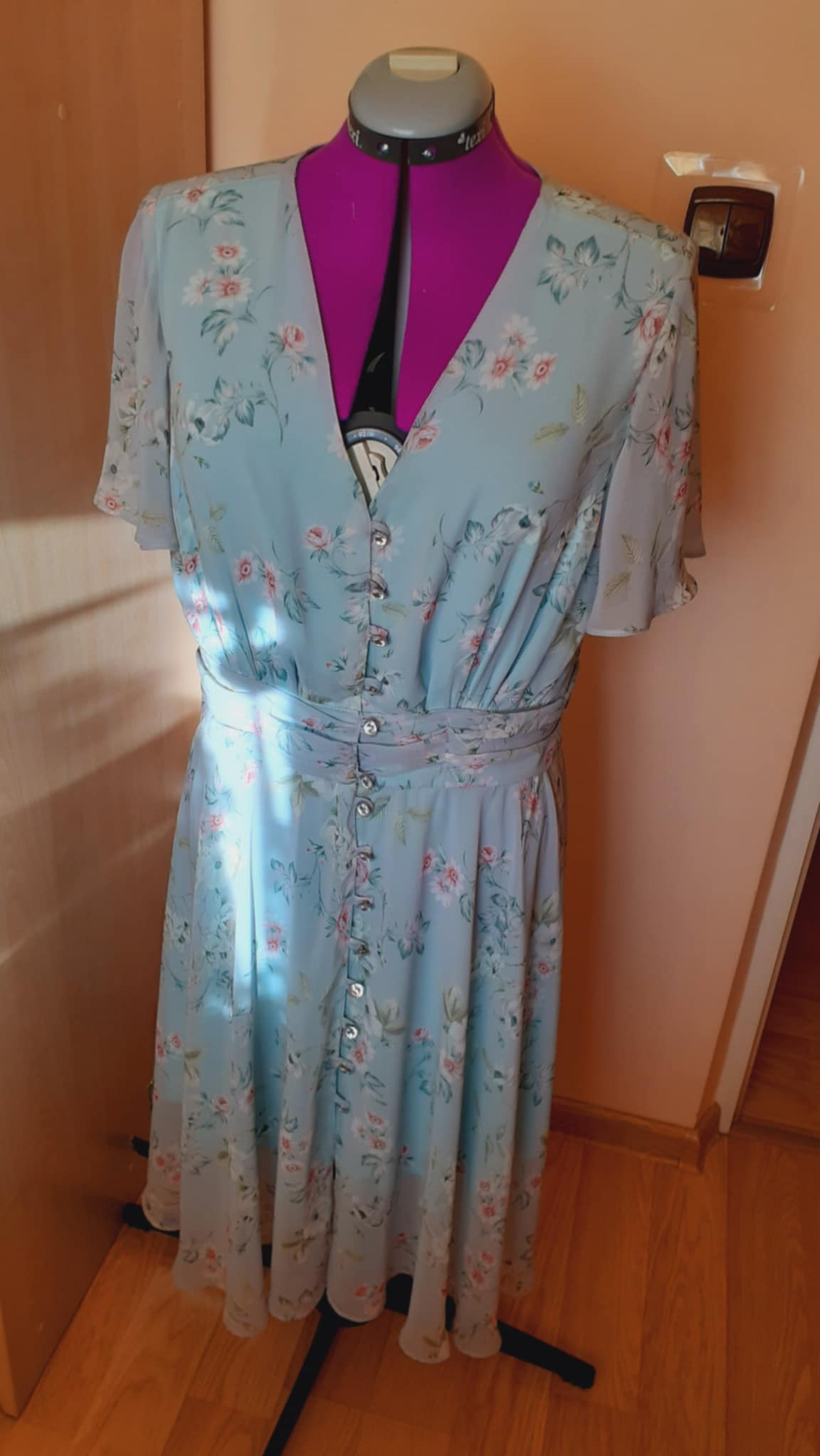 Delikatna, zwiewna sukienka damska w pastelowym błękicie z kwiatowym wzorem, zapinana na ozdobne guziki, prezentowana na manekinie krawieckim z fioletowym korpusem.