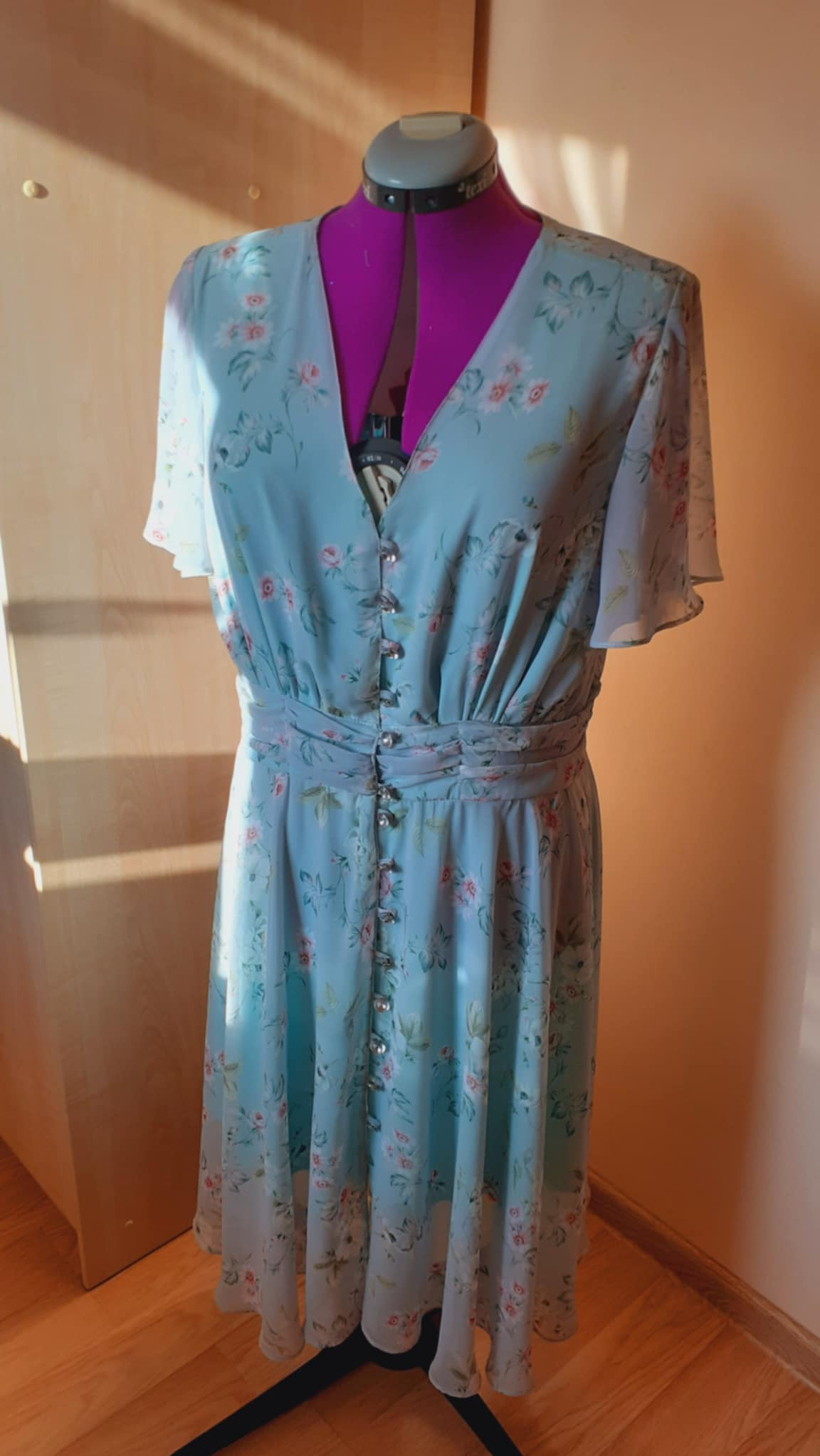 Błękitna sukienka w kwiaty z ozdobnymi guzikami, zapinana na całej długości, prezentowana na manekinie krawieckim z fioletowym korpusem.