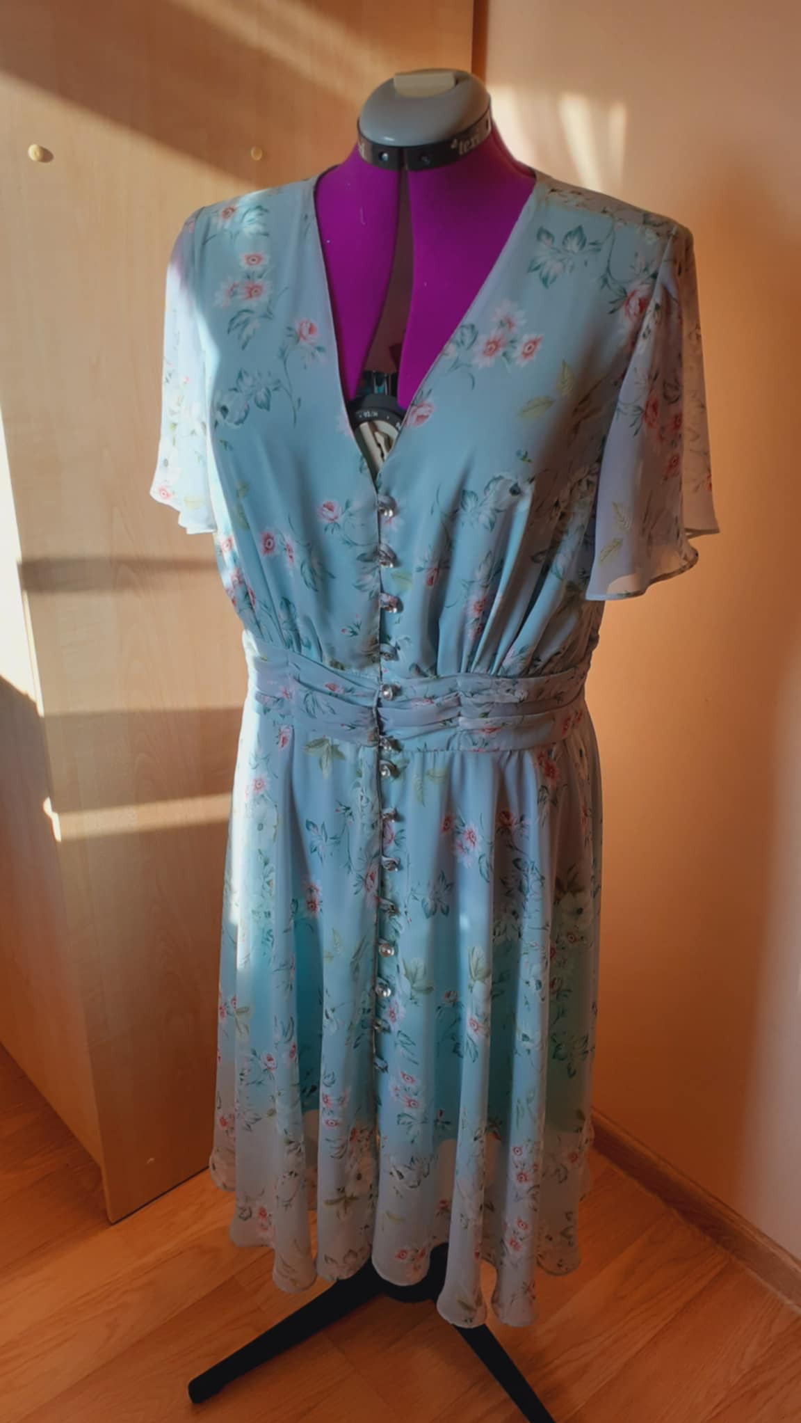 Jasnoniebieska sukienka w kwiaty z ozdobnymi guzikami na manekinie krawieckim z fioletowym torsem.