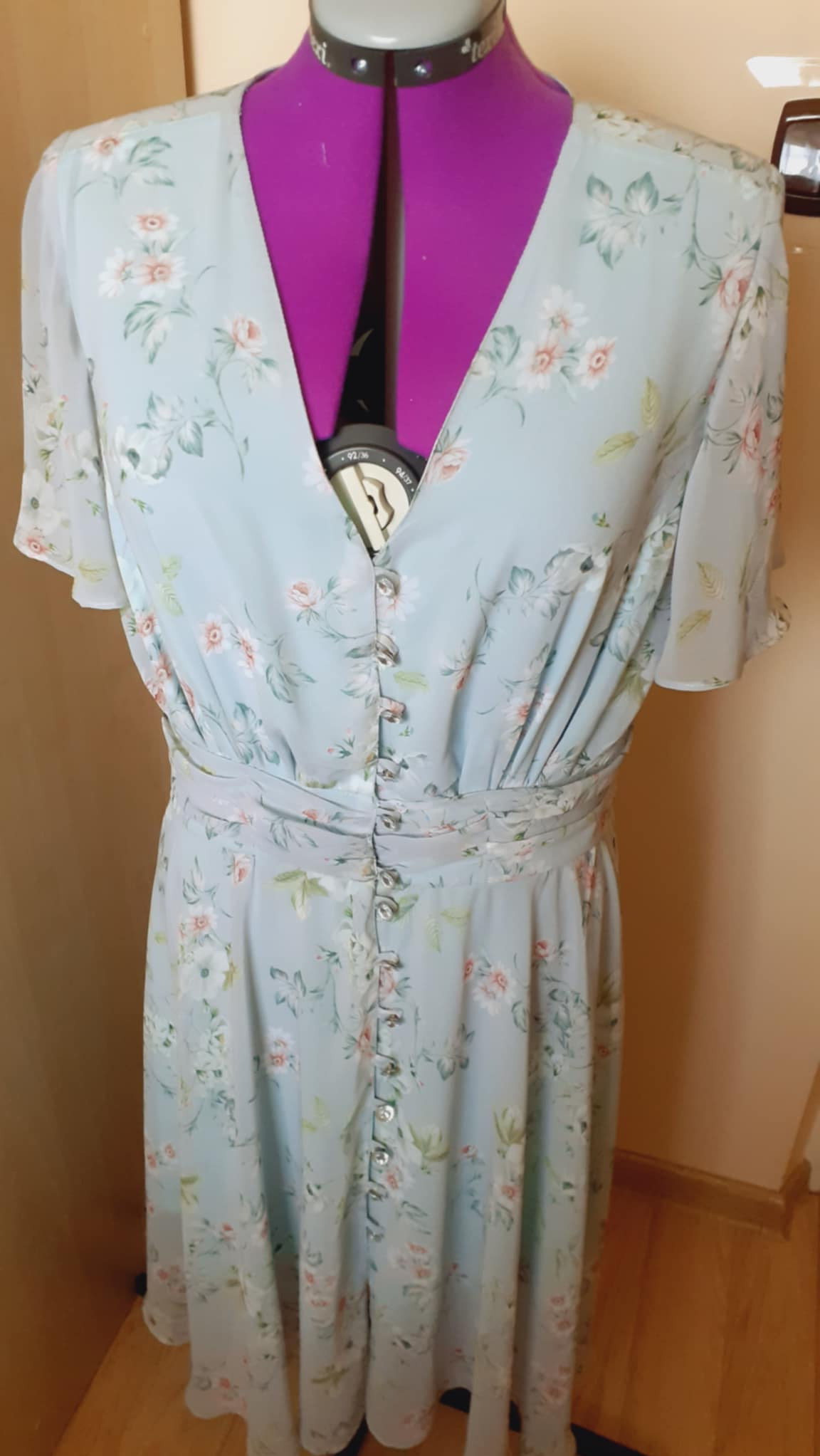 Jasnoniebieska sukienka w kwiaty z ozdobnymi guzikami, umieszczona na różowym manekinie krawieckim.